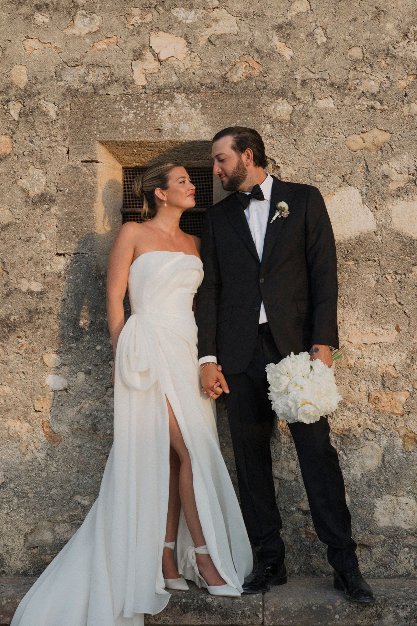 A bride in white gown and groom in tuxedo share intimate moments against a rustic stone wall backdrop.