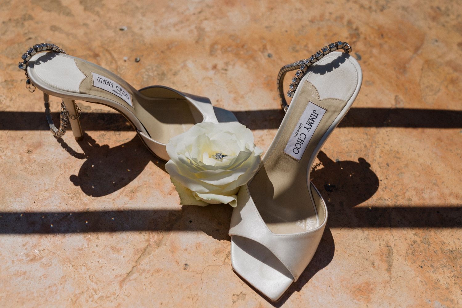 White Jimmy Choo bridal heels with crystal ankle straps and rose detail displayed on marble floor.