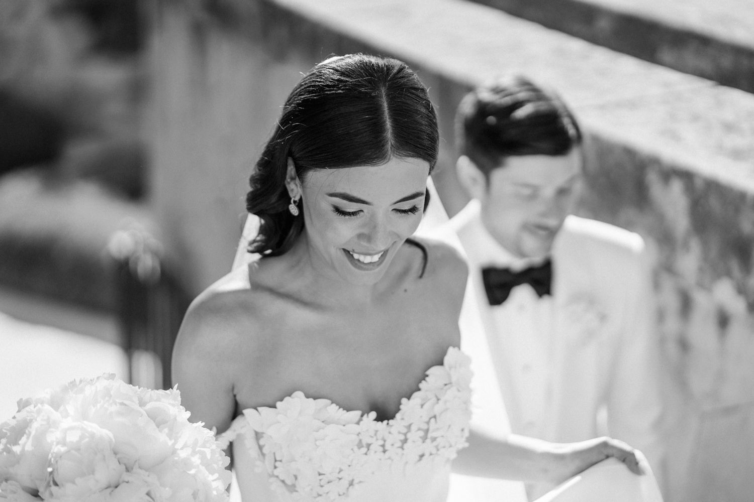 Black and white wedding photo of smiling bride in strapless gown holding bouquet with groom in tuxedo behind her.