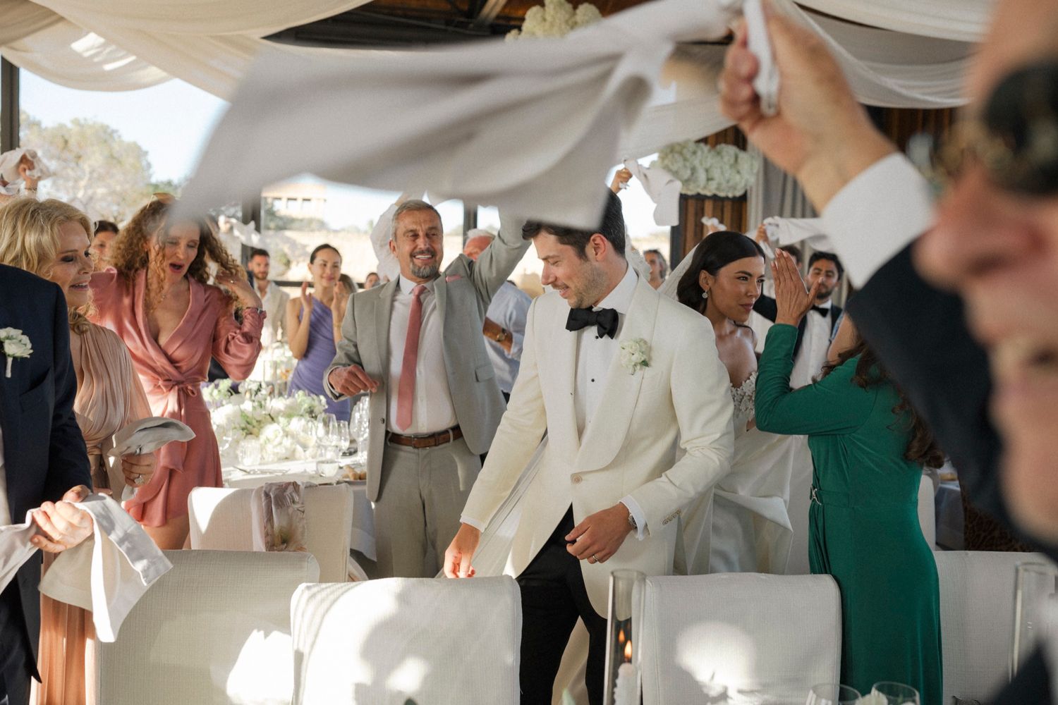 Groom in white tuxedo celebrated by wedding guests waving white napkins at elegant outdoor reception.