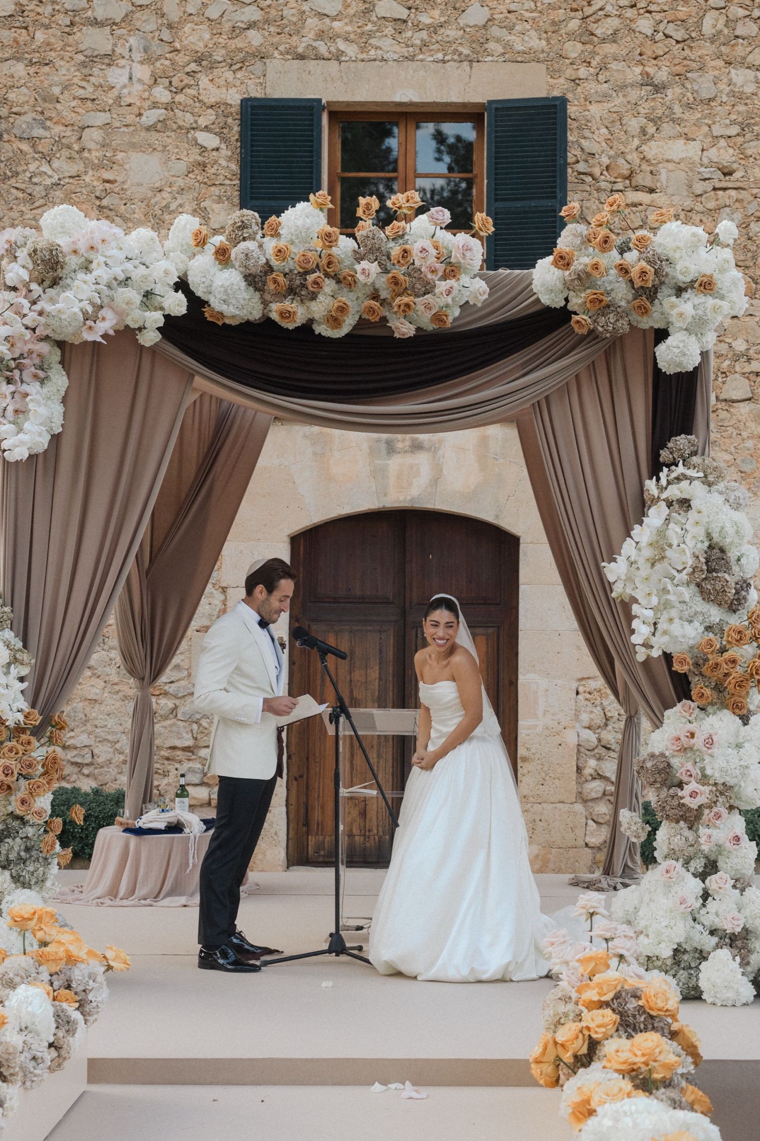 Elegant wedding ceremony setup with stone walls, beige drapes, and white floral arrangements creating a romantic atmosphere.
