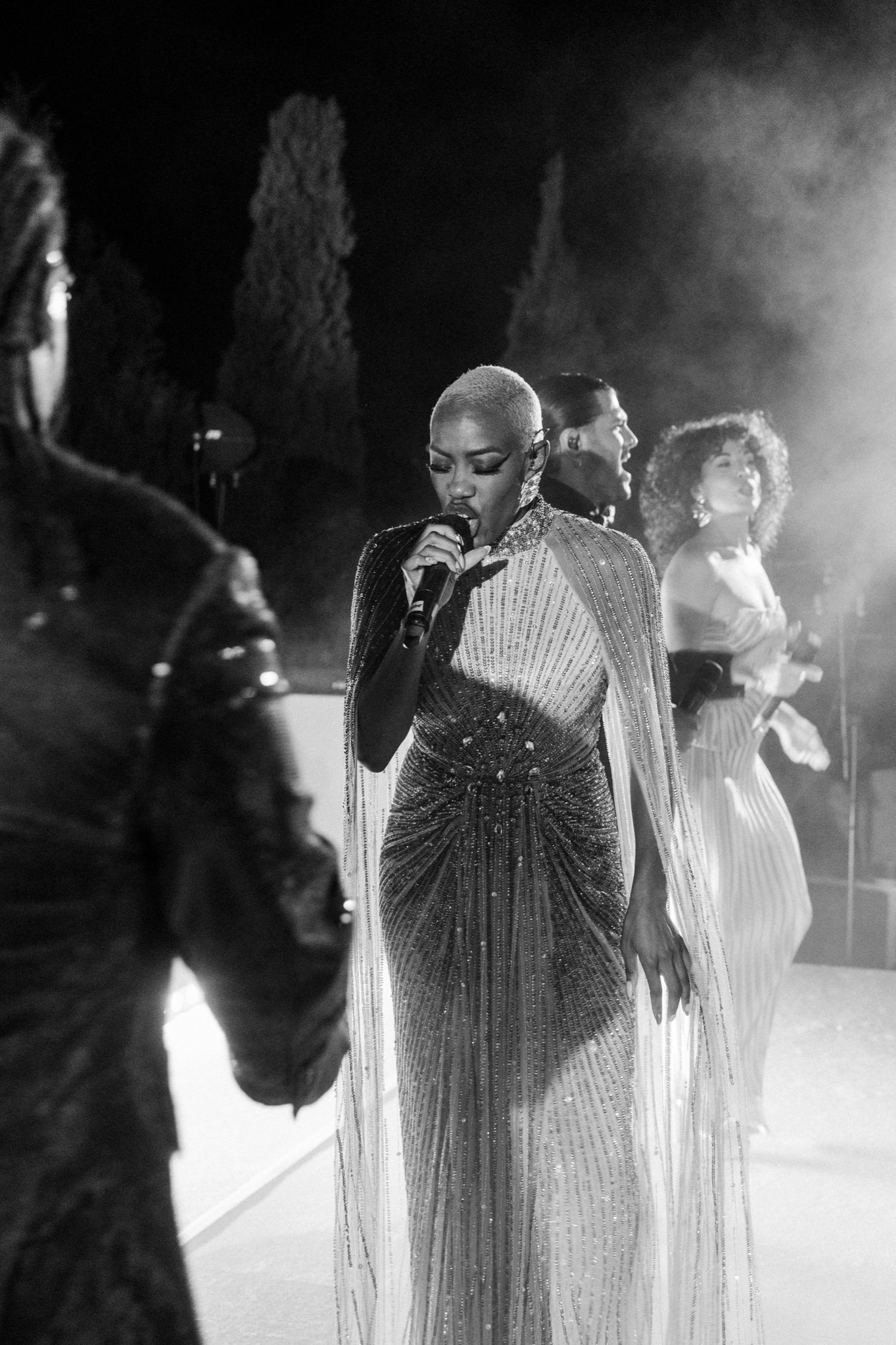 Performers in sparkly dresses sing on stage with dramatic backlighting and fog effects.