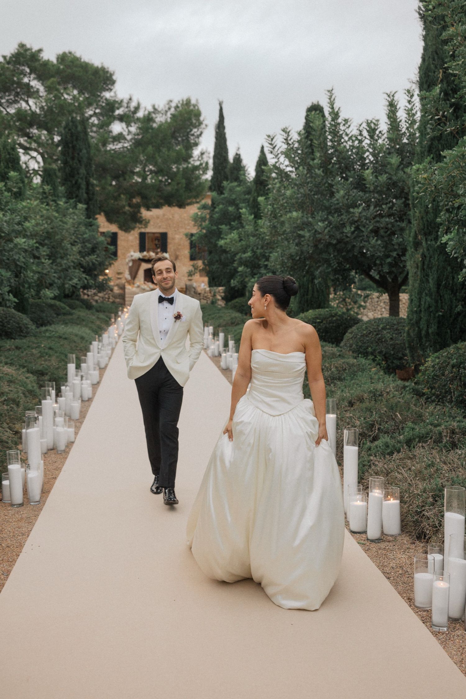 A couple in wedding attire walks down a path lined with glowing candles in glass holders at dusk.