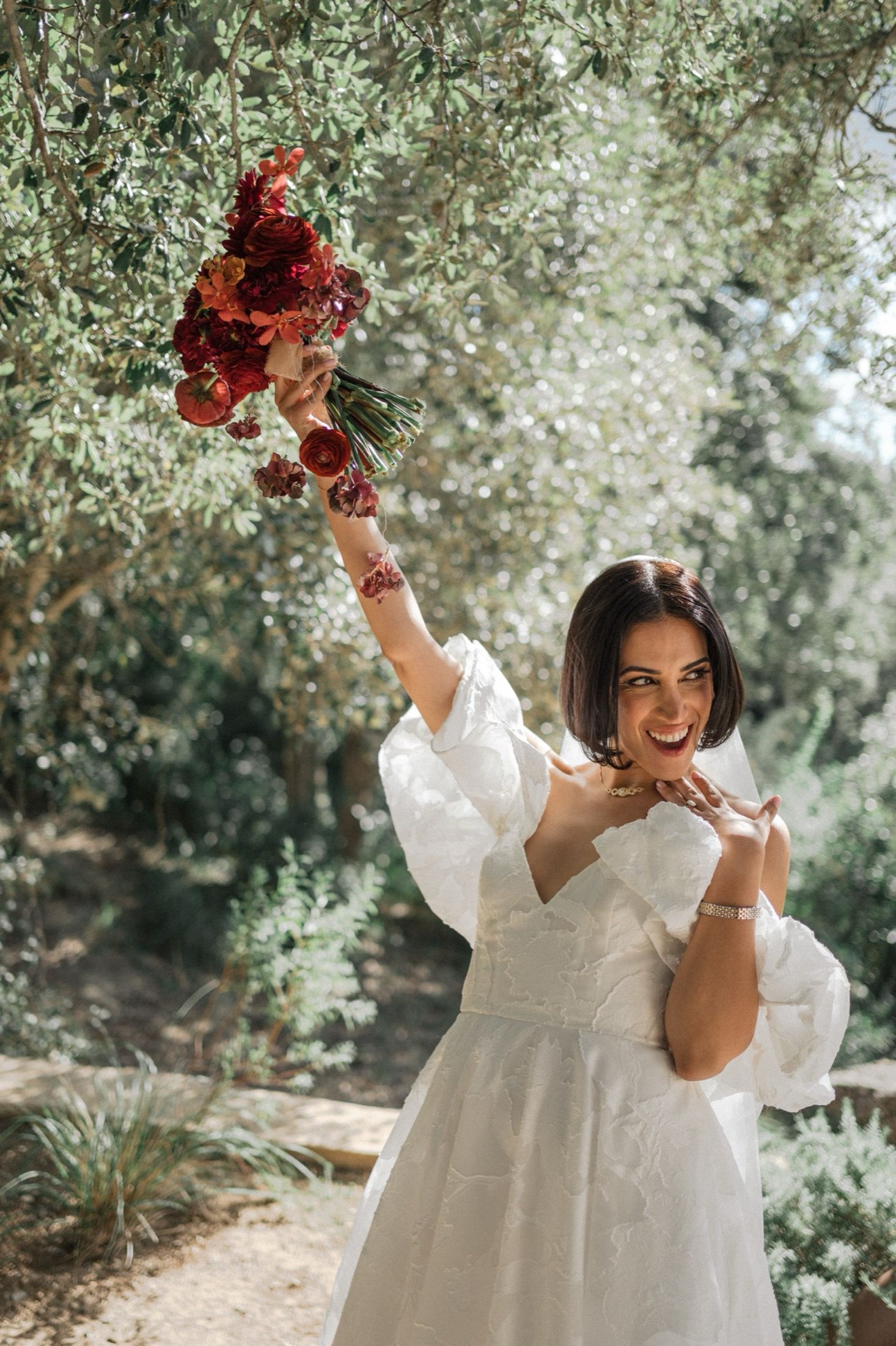 A person in a white dress holds up a bouquet of deep red flowers in a sunlit garden setting.