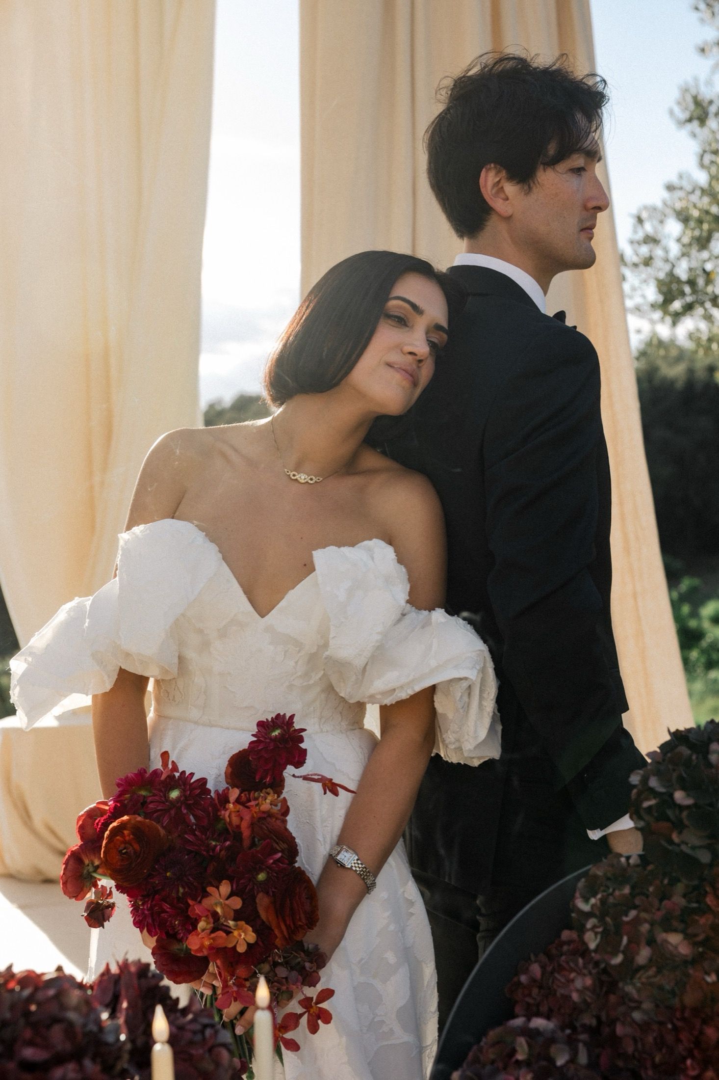Romantic wedding couple poses in white dress and black suit with deep red bouquet against sunlit columns.