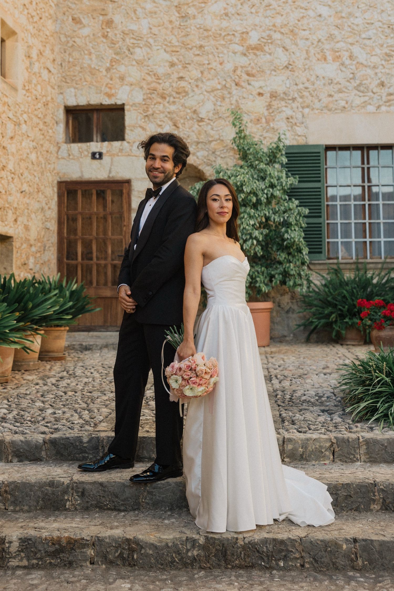 A wedding couple poses on stone steps against a rustic Mediterranean building with green plants and terracotta walls.