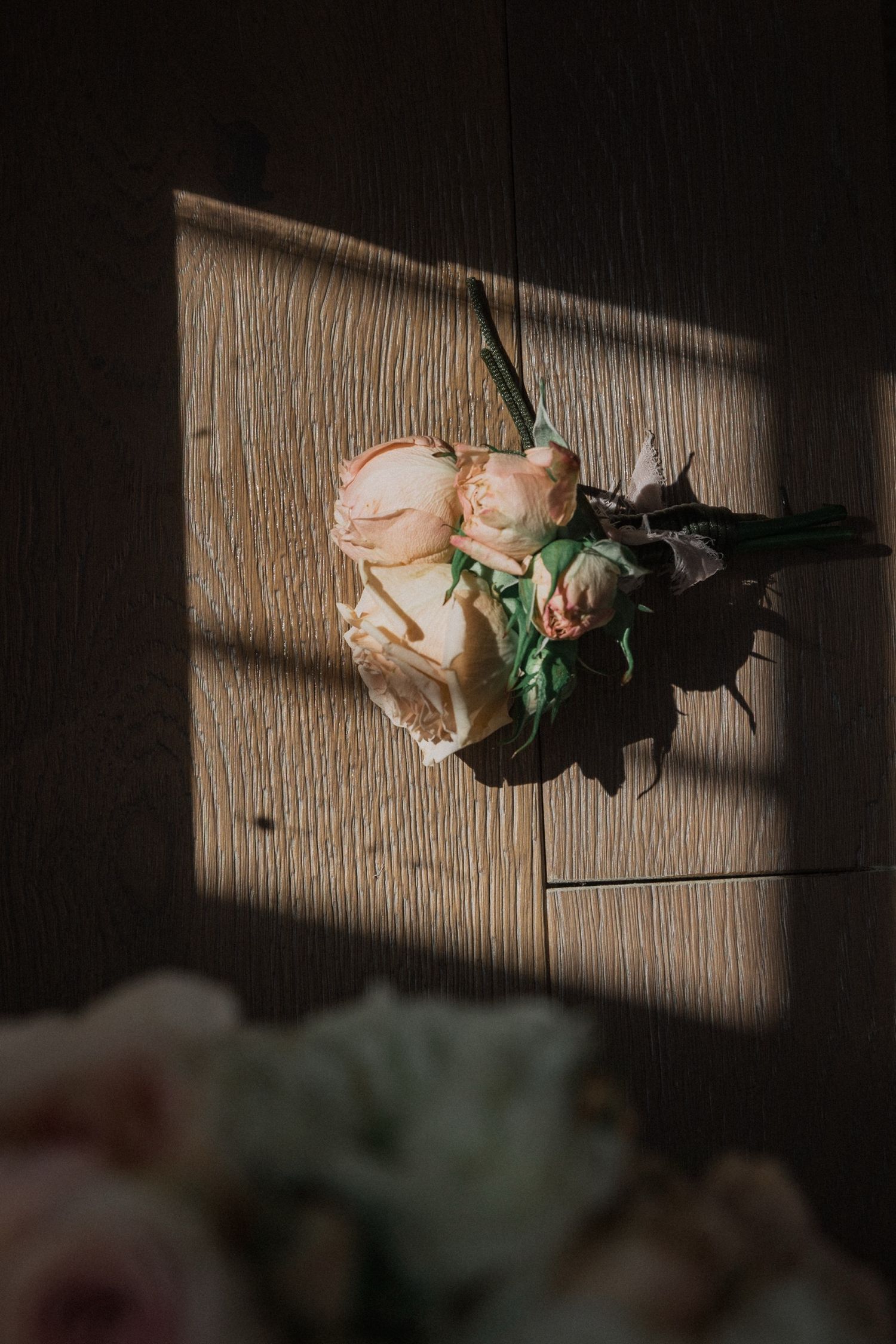 A pink rose with green leaves casts dramatic shadows on a textured wooden surface in moody lighting.
