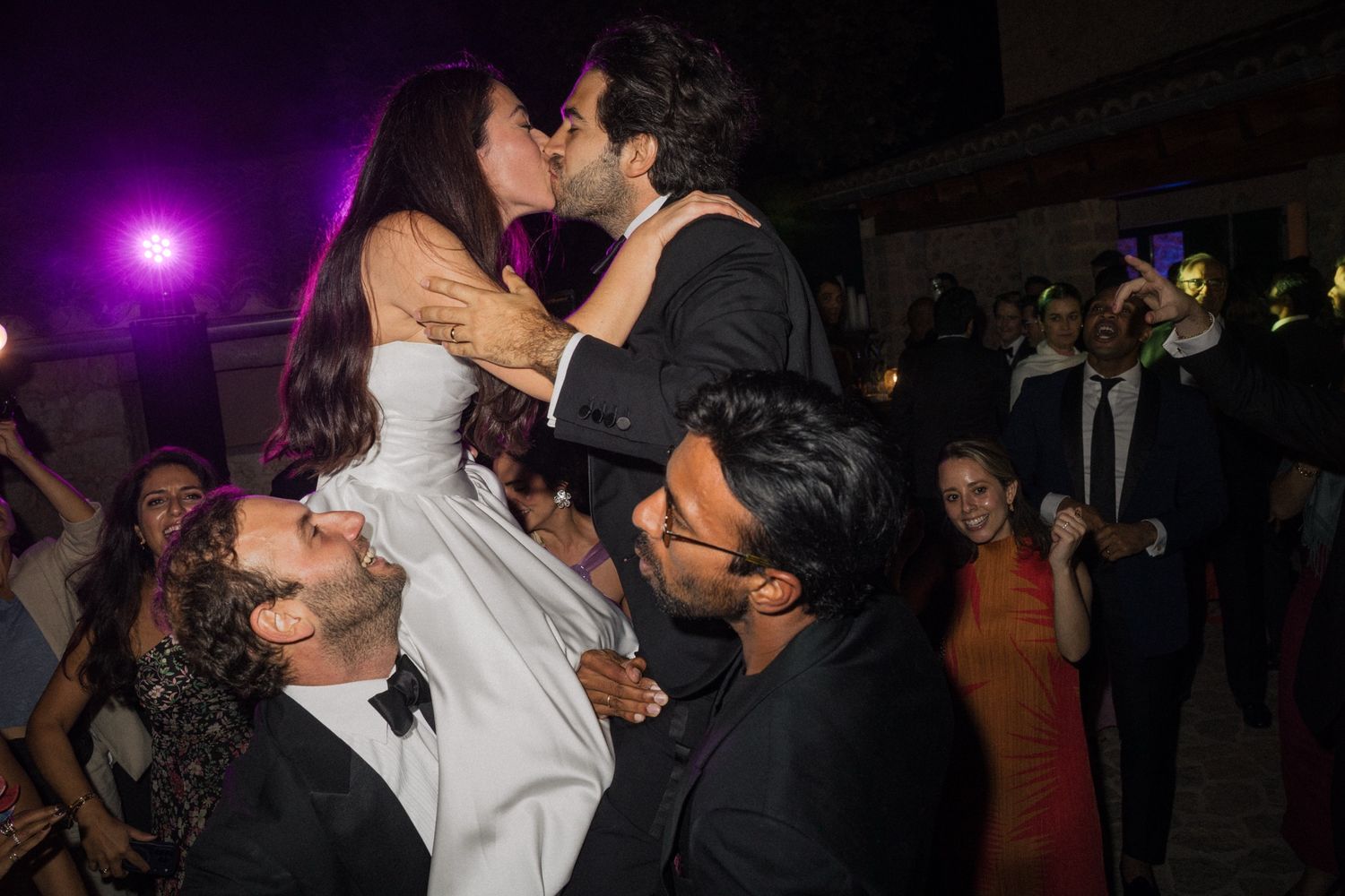 A couple shares a romantic moment at a wedding reception while being lifted by guests on the dance floor under purple lighting.