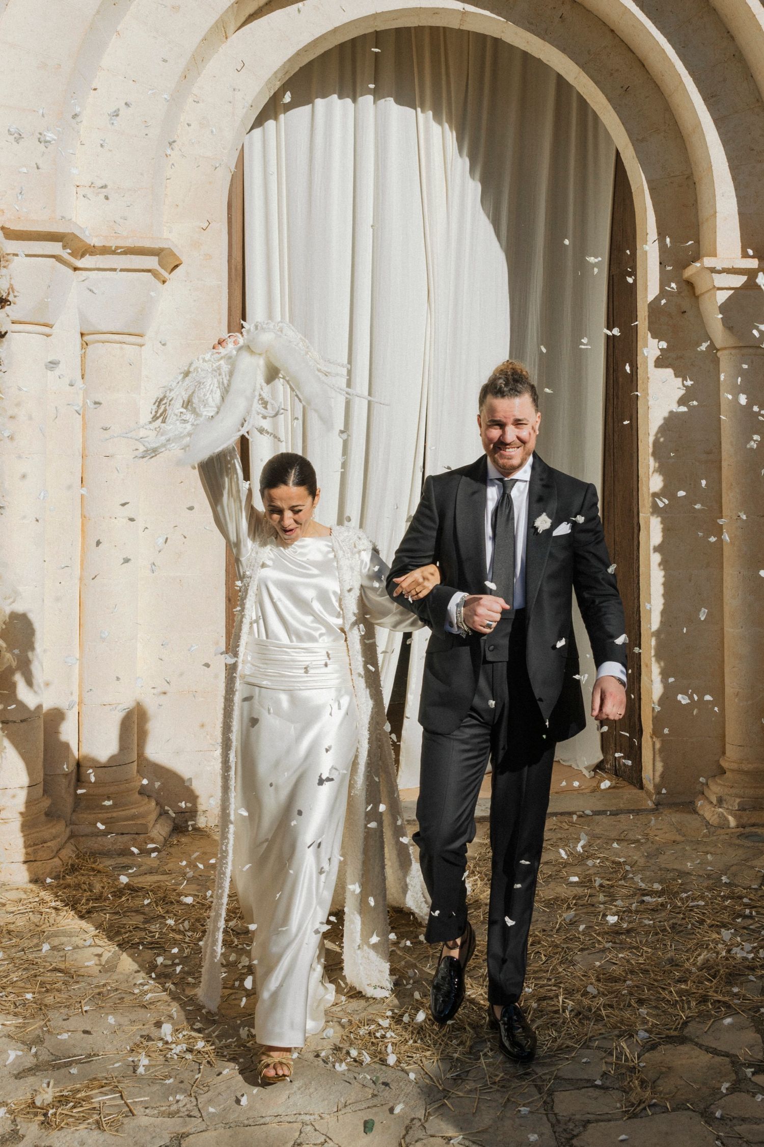 Wedding couple walking through stone archway with confetti falling in natural light.