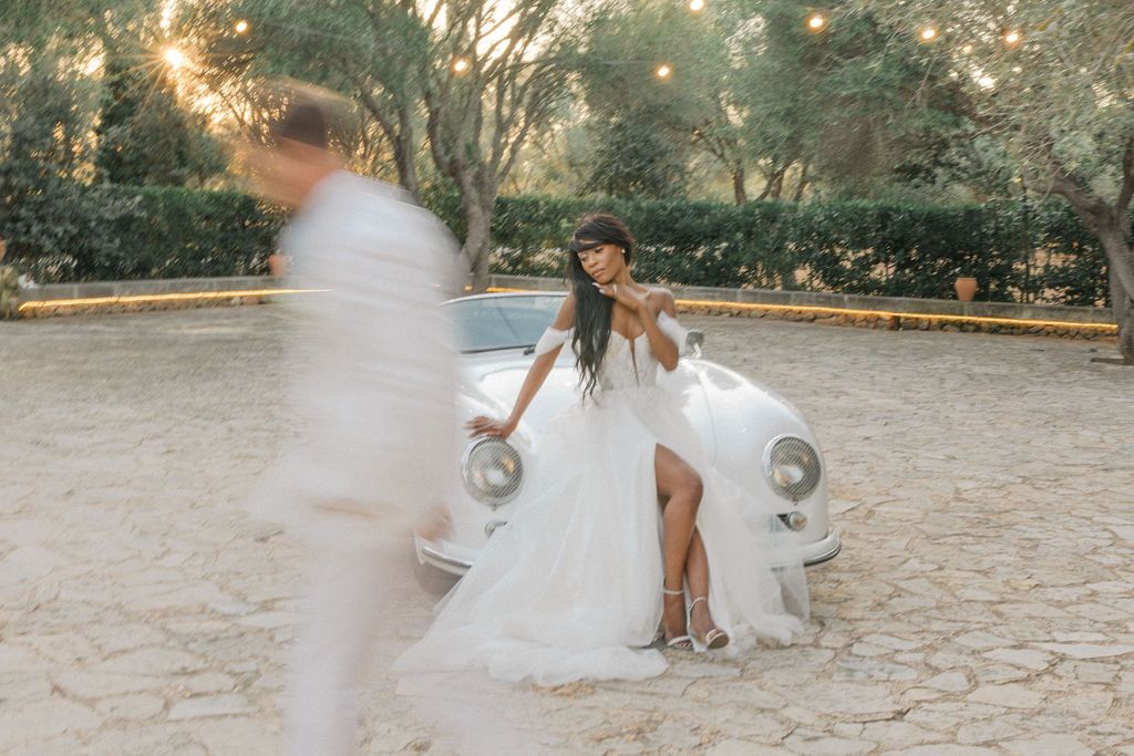 A romantic scene with a vintage car and flowing white dress in a garden setting.