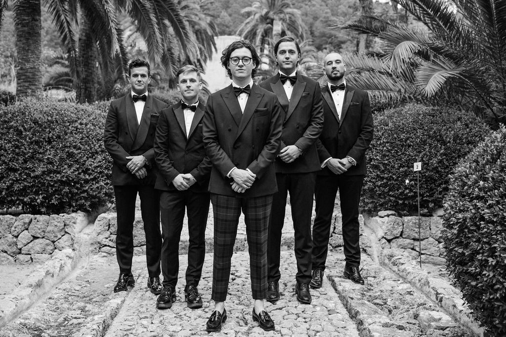 Groomsmen in black tuxedos pose together on stone steps in garden setting.