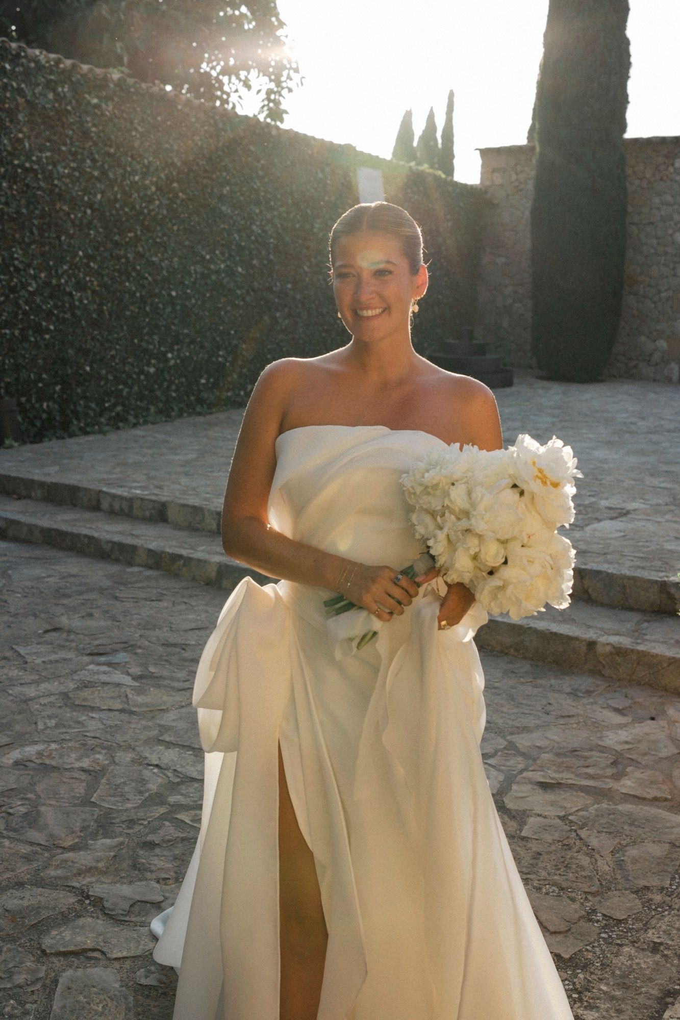 Bride holding white bouquet poses in flowing off-shoulder gown against stone wall.