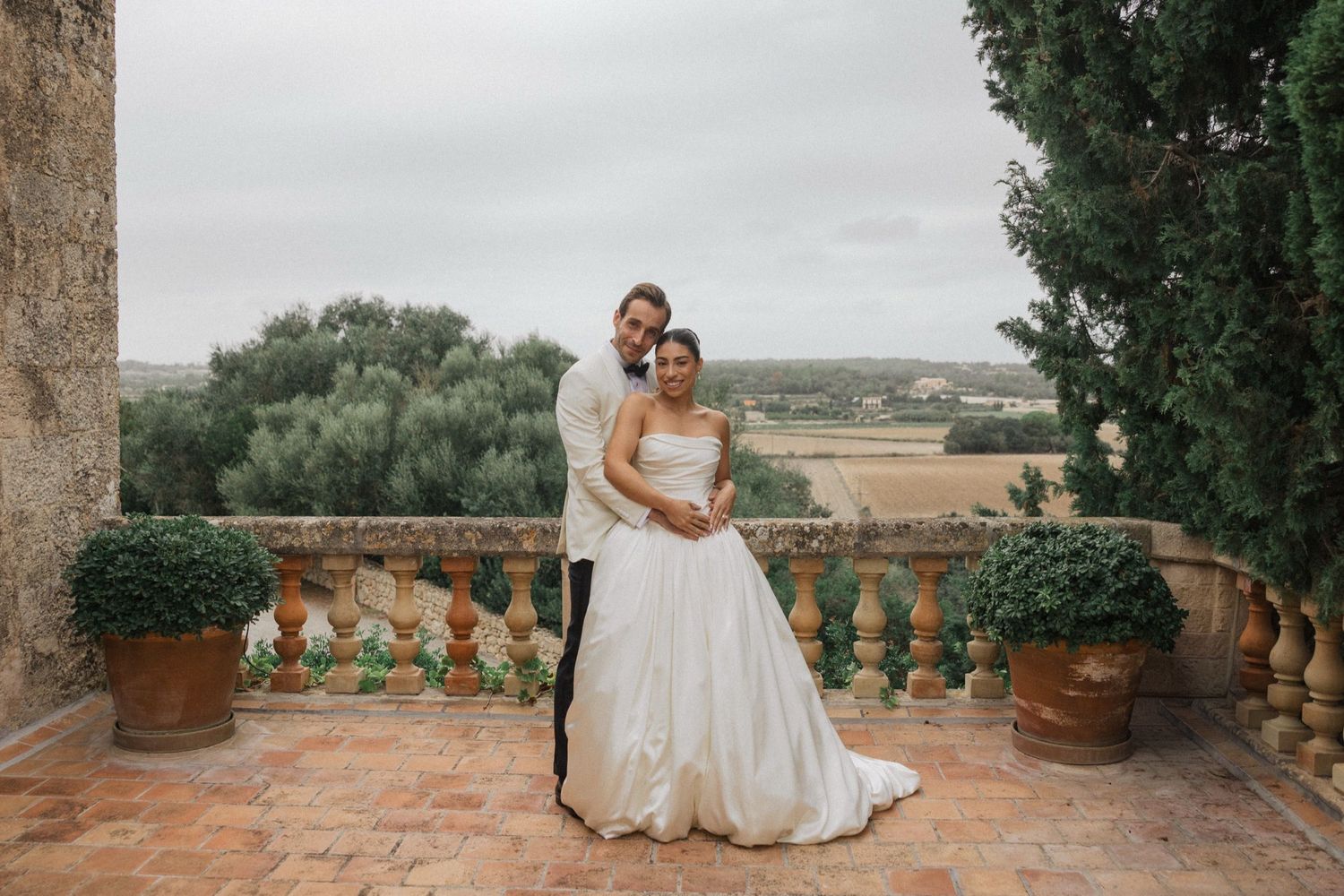 A romantic wedding portrait of a couple embracing on a Mediterranean-style terrace with potted plants and scenic views.