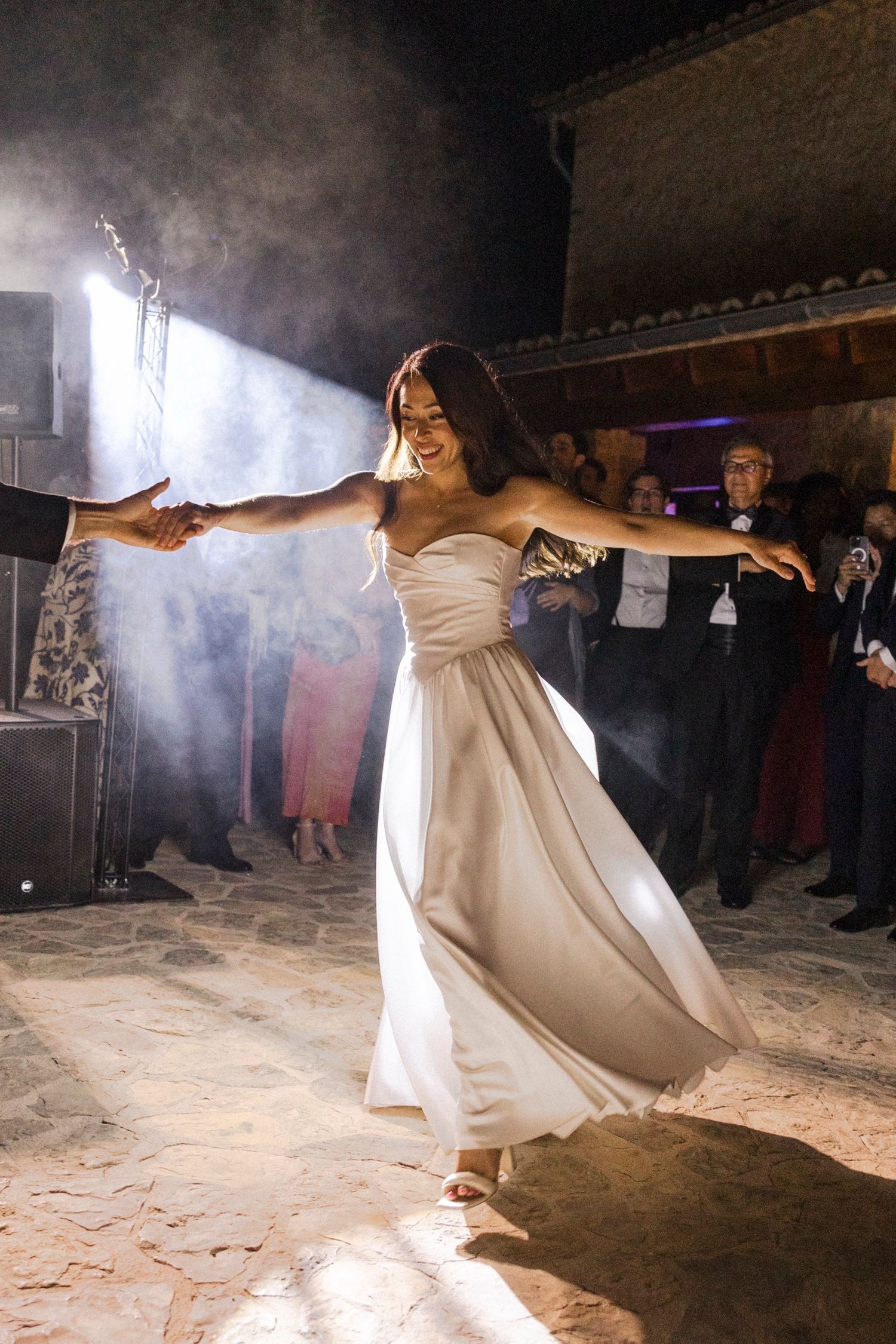 A bride in a flowing white gown dances joyfully in a beam of light while guests watch in the background.