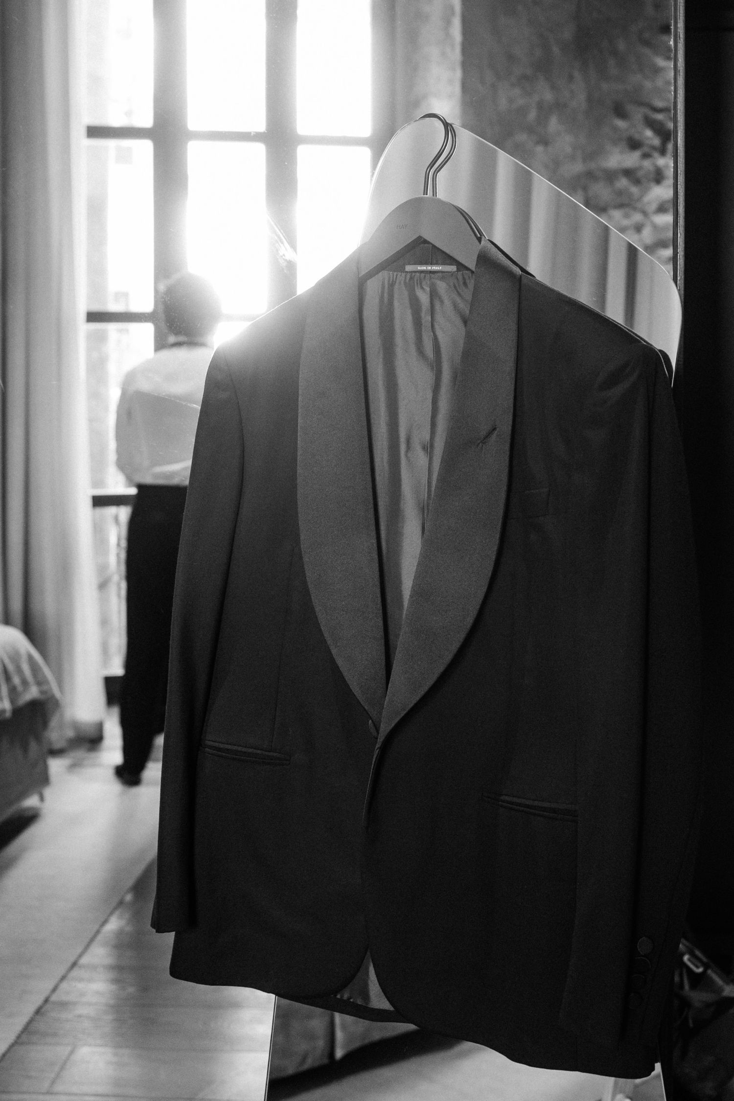 A black tuxedo jacket with shawl lapels hangs on a hanger near a sunlit window in black and white photography.