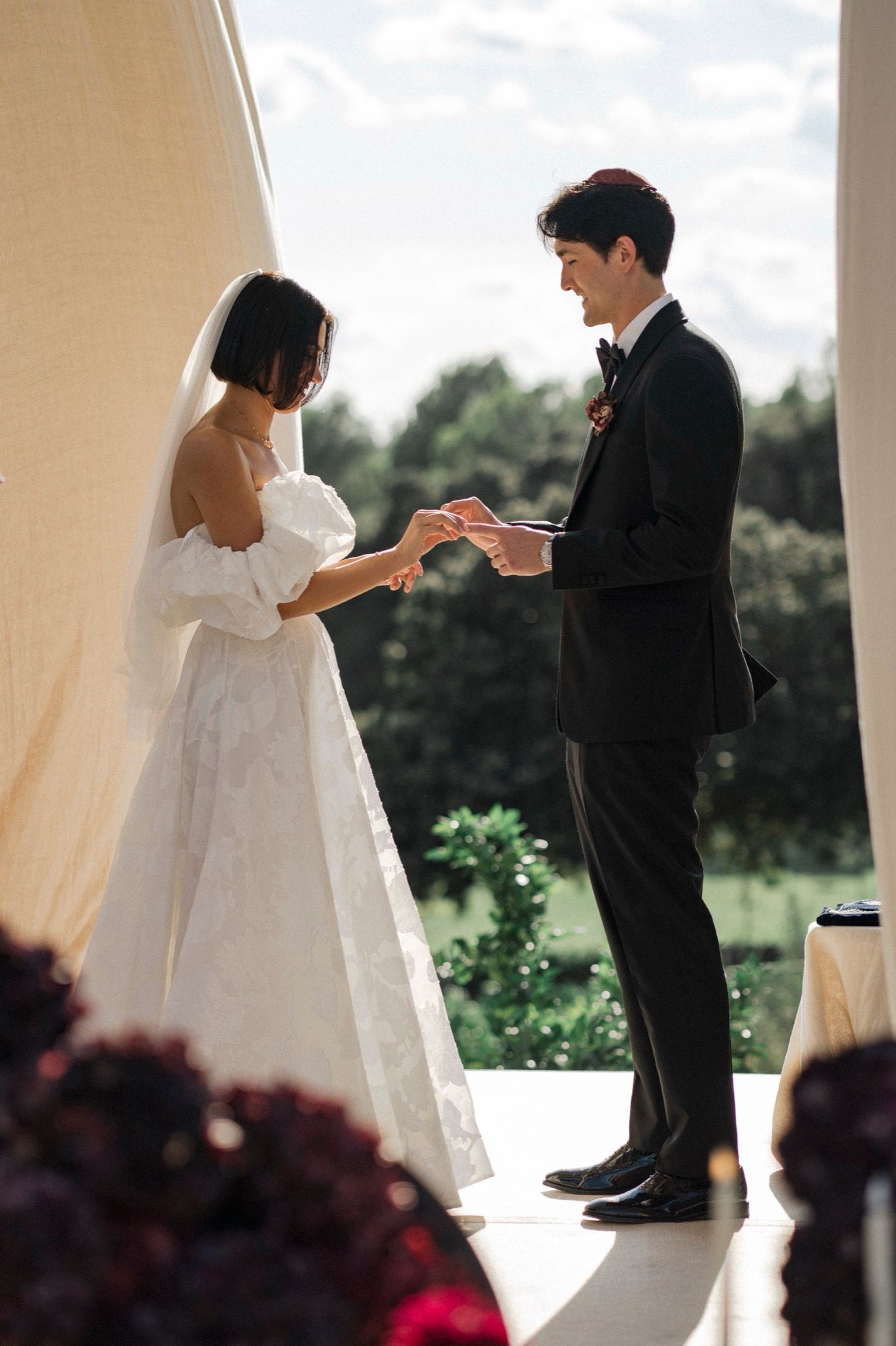 A couple exchanges vows during their wedding ceremony under a white draped archway outdoors.