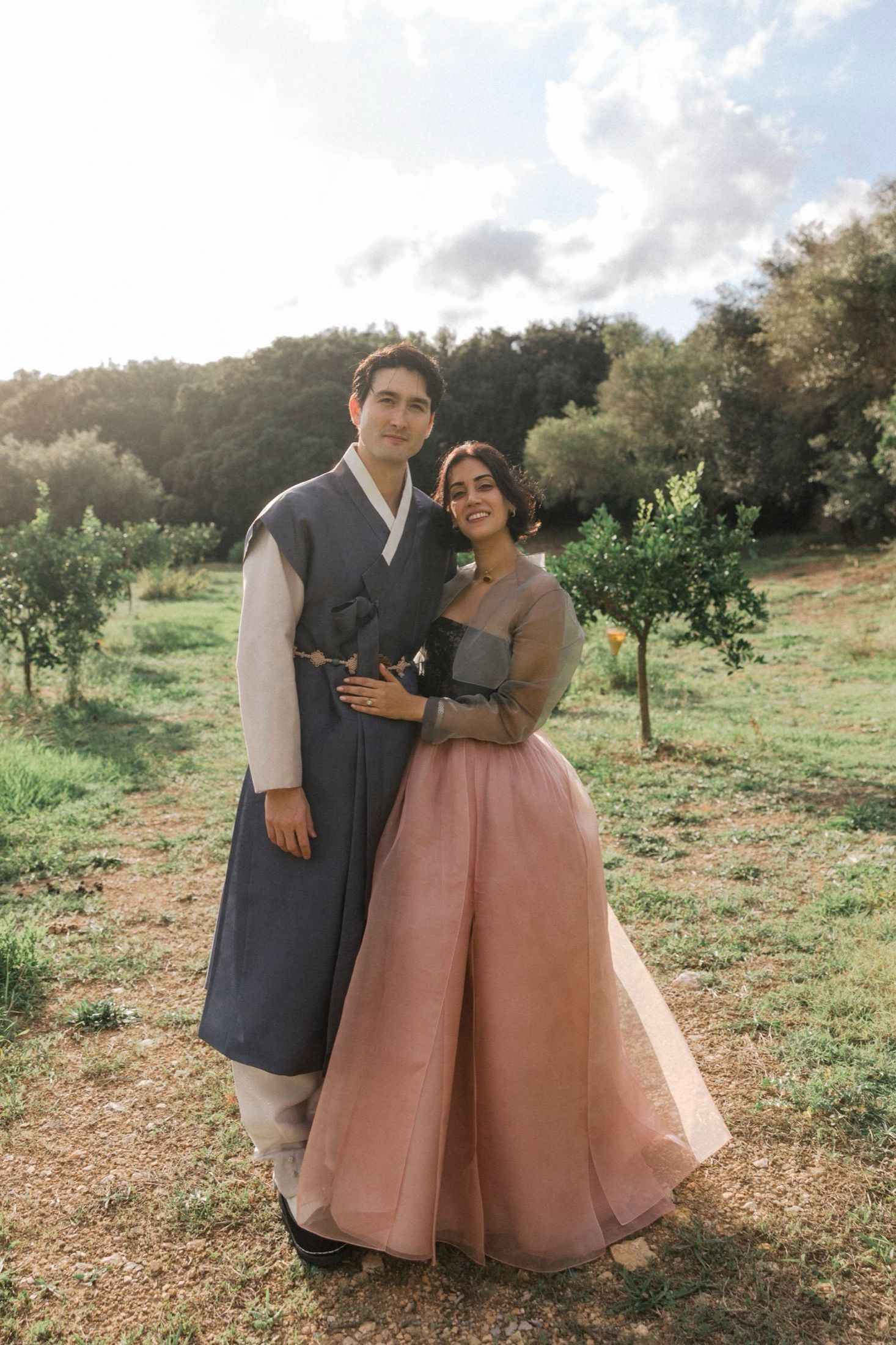A couple in formal attire embraces while standing in a sunny orchard with trees in the background.