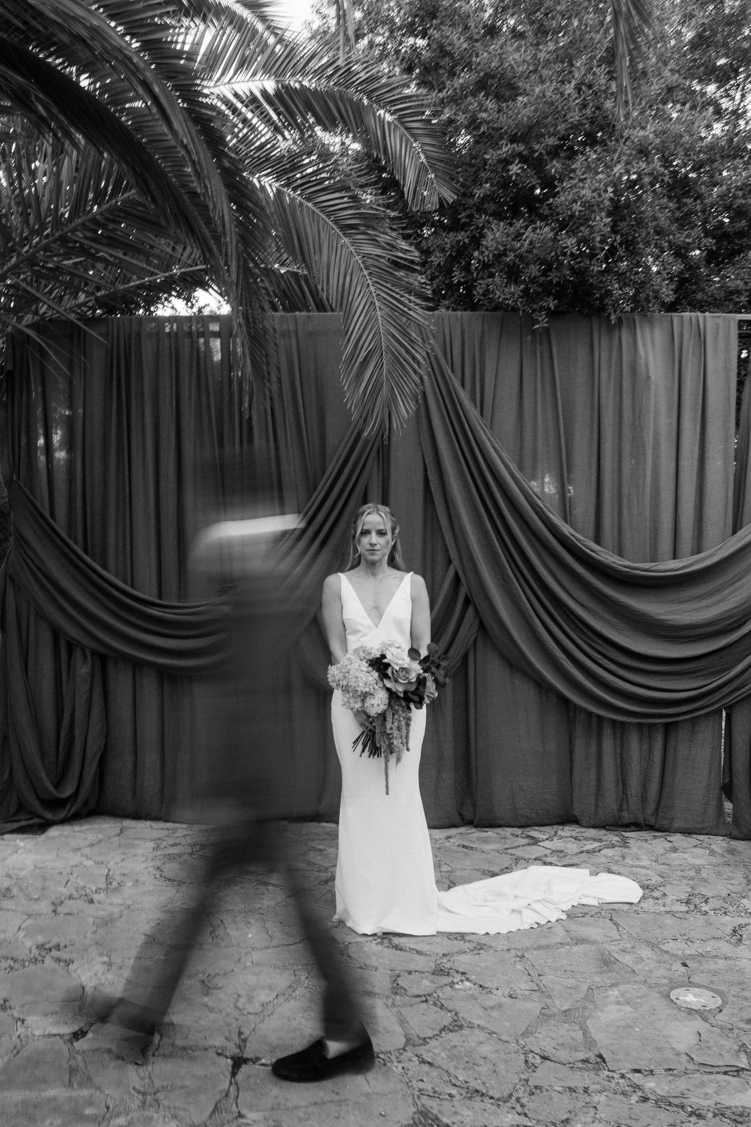 Black and white wedding photo showing a person in a long white dress holding a bouquet under palm fronds and draped fabric.