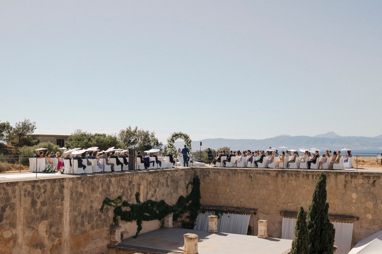 Outdoor wedding ceremony on a stone terrace overlooking the Mediterranean sea with guests seated in white chairs.