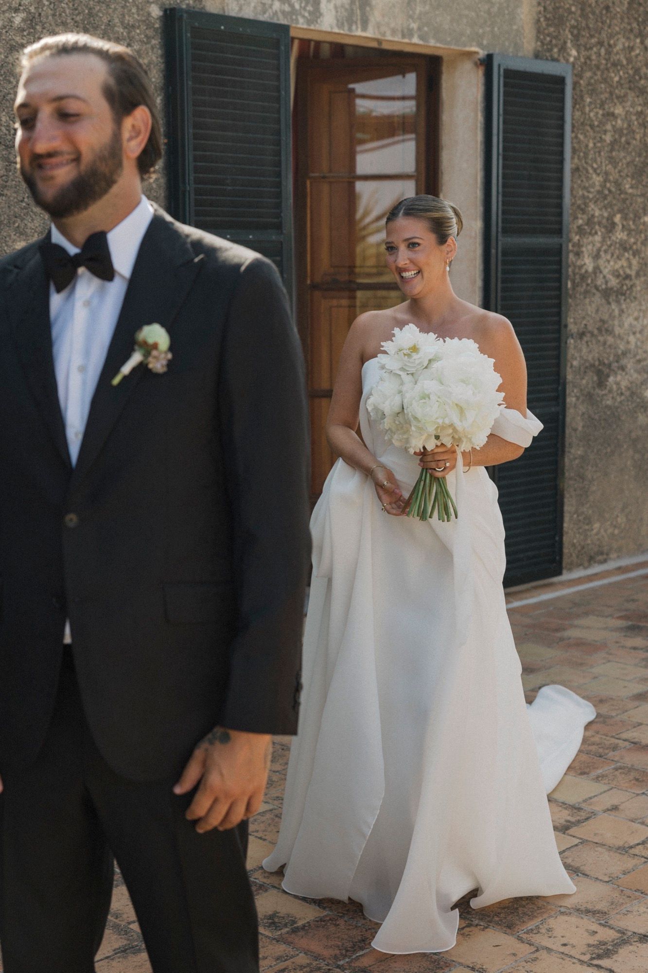 Bride and groom share a moment on a stone terrace with rustic shuttered windows.