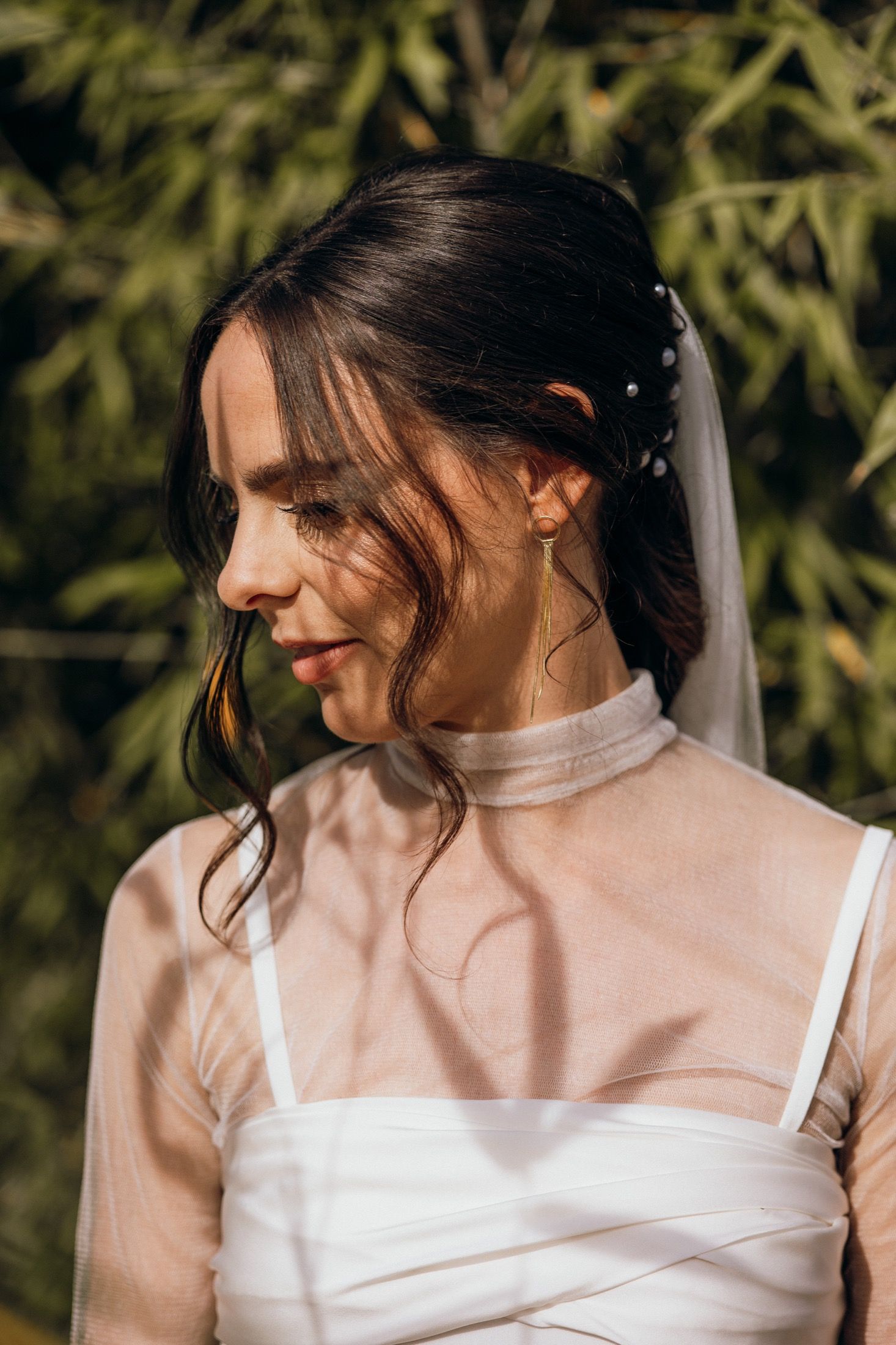 A bride in a white wedding dress poses outdoors with soft natural lighting and loose tendrils of dark hair framing her face.