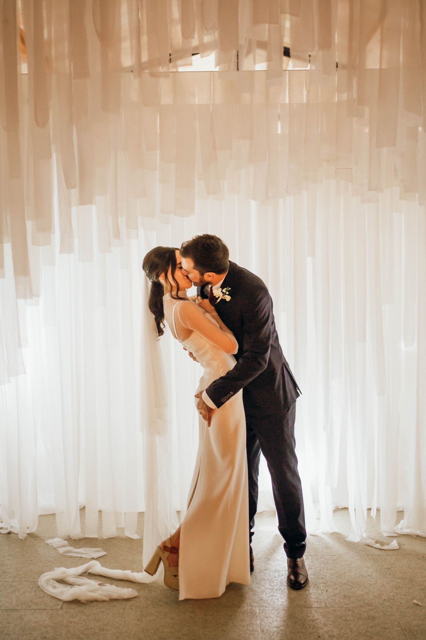 A romantic wedding moment with a couple sharing a kiss against a backdrop of flowing white curtains.