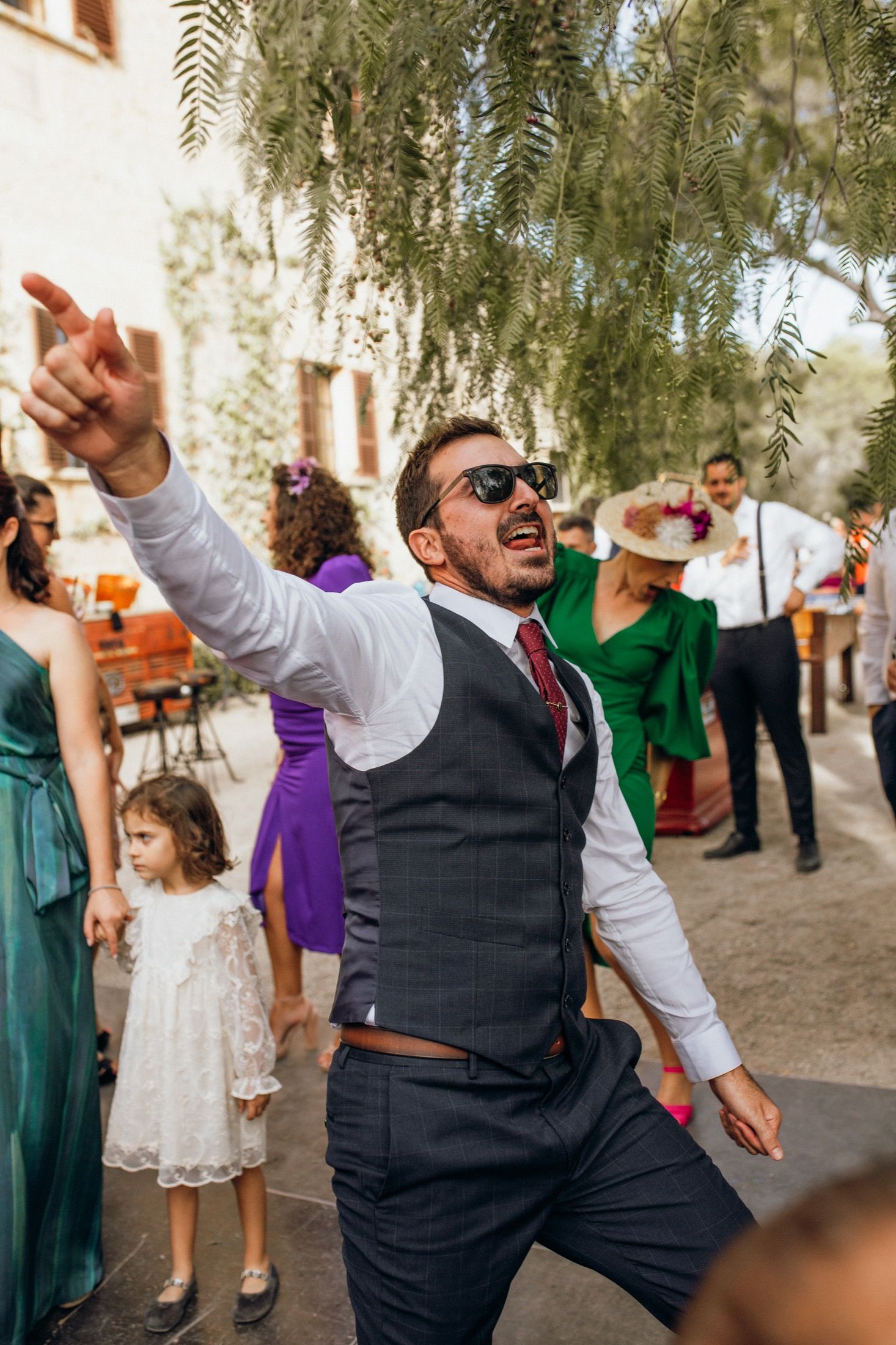Wedding guests dancing and celebrating joyfully at an outdoor reception under willow trees.