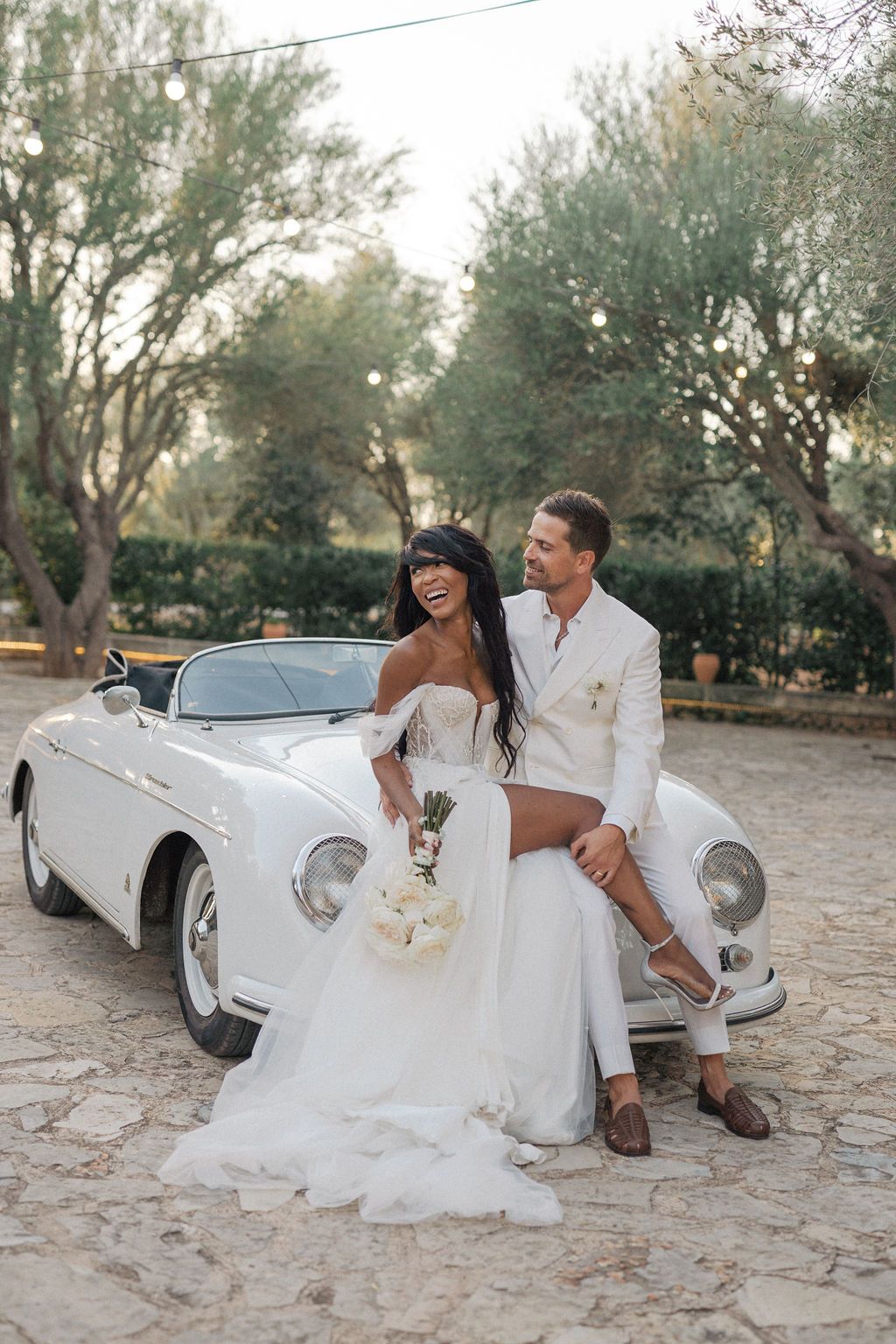Wedding couple laughing together beside vintage convertible car.
