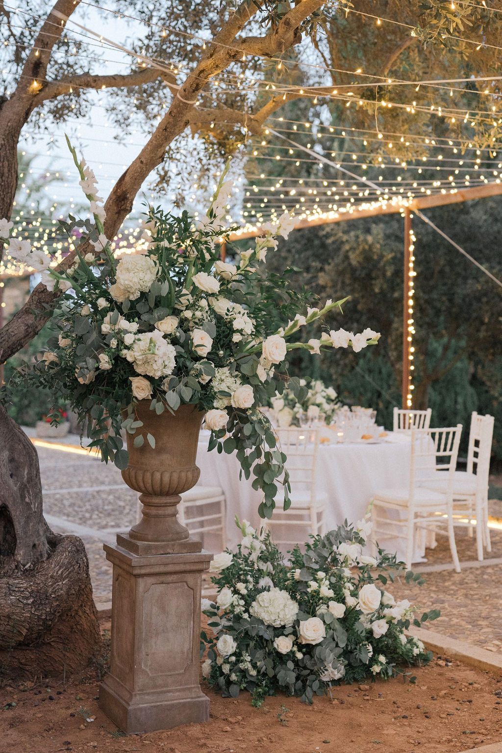 Elegant outdoor wedding decor with white floral arrangements in stone urns under string lights and trees.