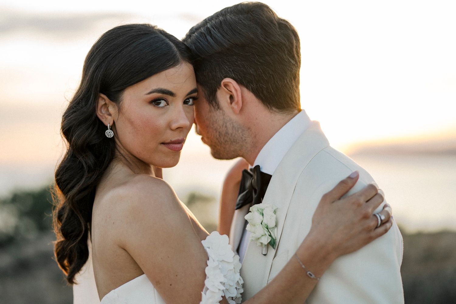 Bride and groom share an intimate moment at sunset, with the groom in a white suit and bride in a strapless gown.