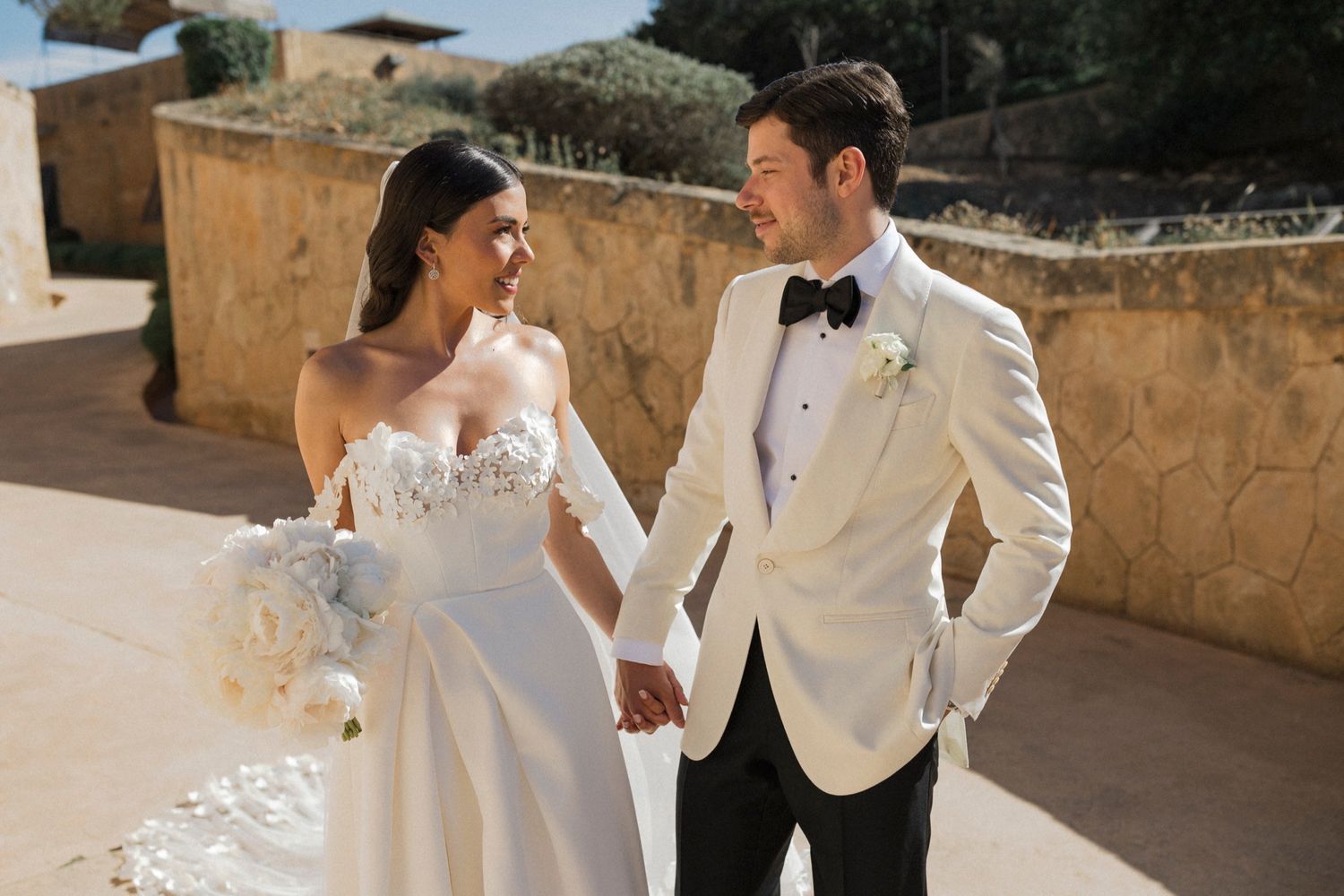 Bride and groom holding hands smiling at each other outdoors, wearing elegant white wedding attire with bouquet.