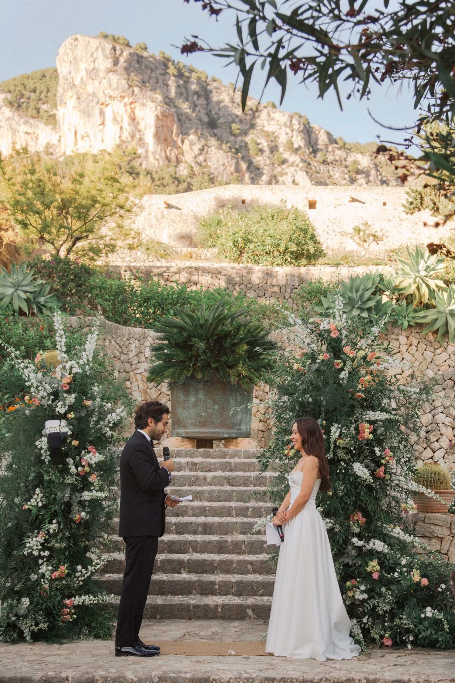 A wedding ceremony unfolds on rustic stone steps adorned with floral arrangements and greenery against a mountainous backdrop.
