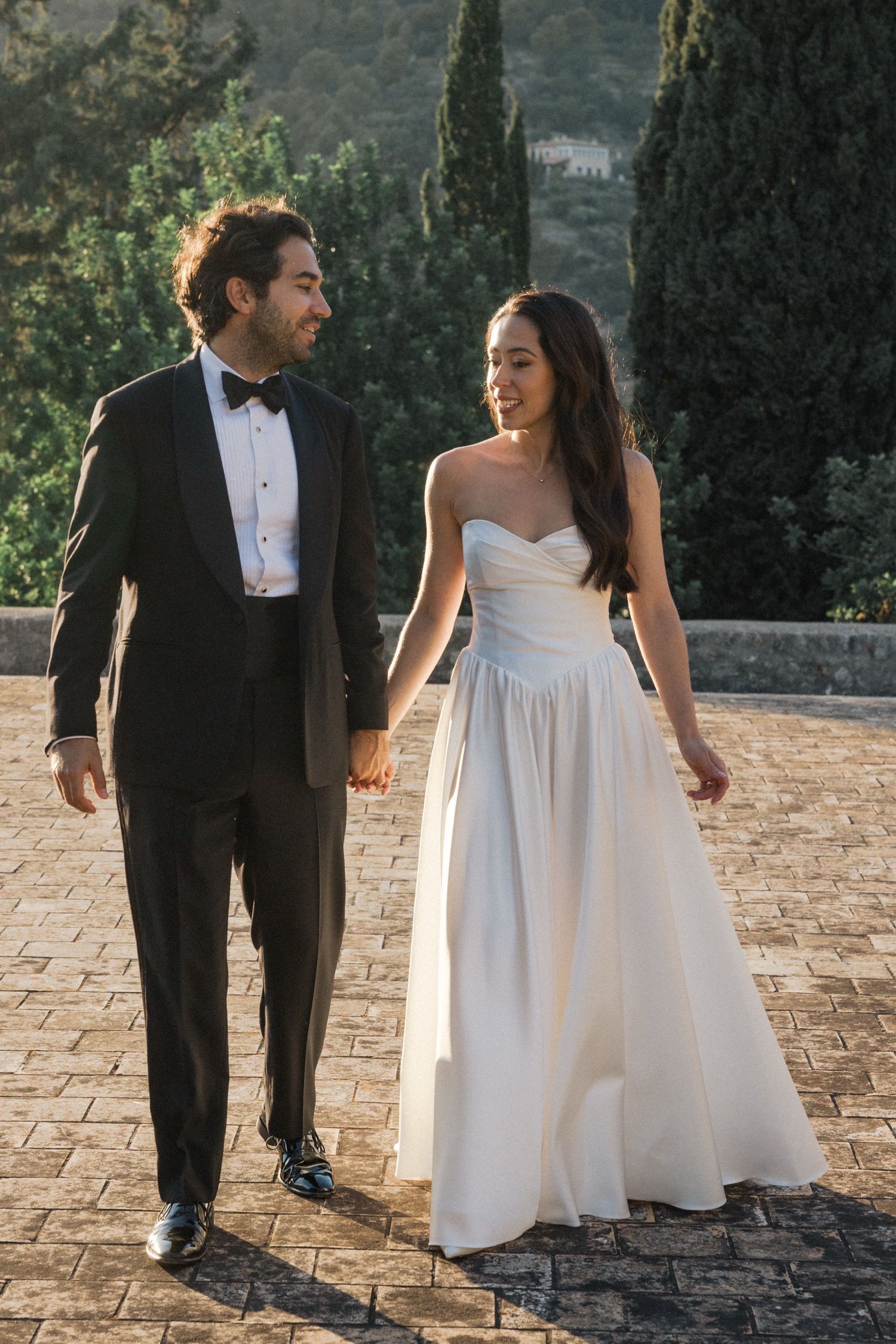 A couple in wedding attire walks along a stone path surrounded by cypress trees in a romantic outdoor setting.