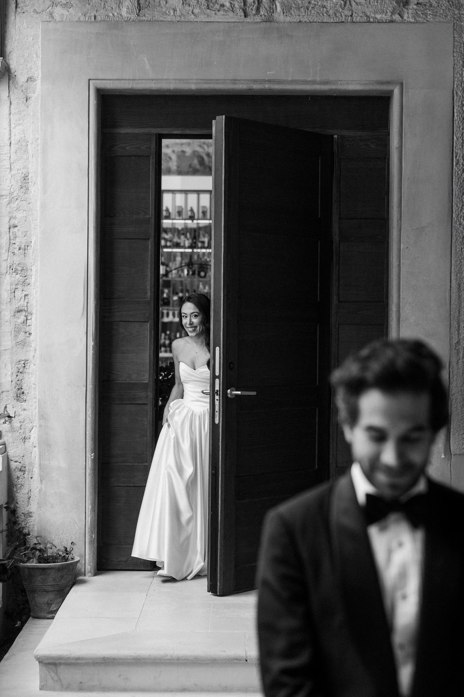 A bride in a white gown stands in a doorway while a groom in a tuxedo is positioned in the foreground of this black and white photo.
