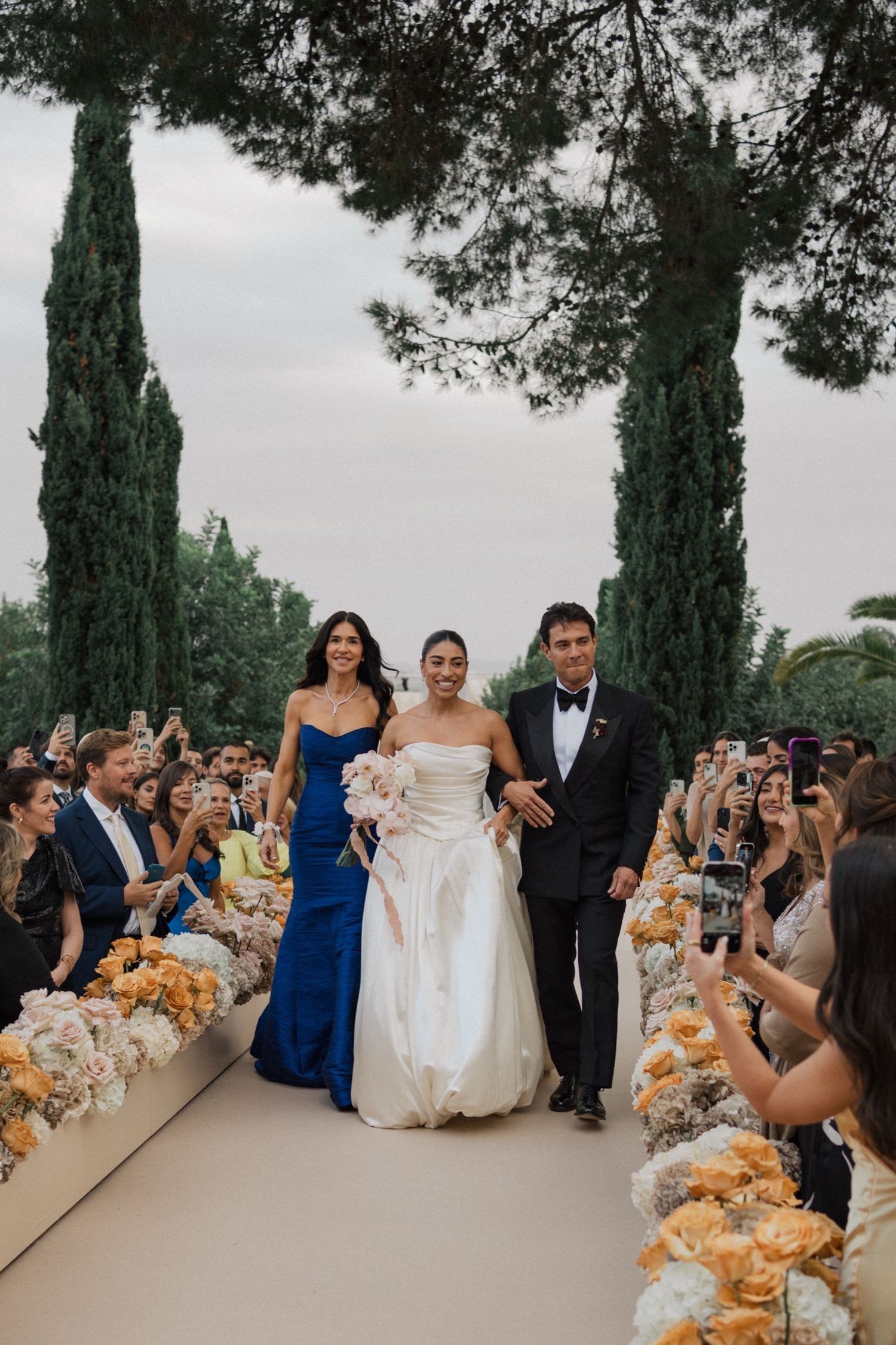 Wedding guests capture photos on phones as newlyweds walk past elegant outdoor cypress tree ceremony setup.