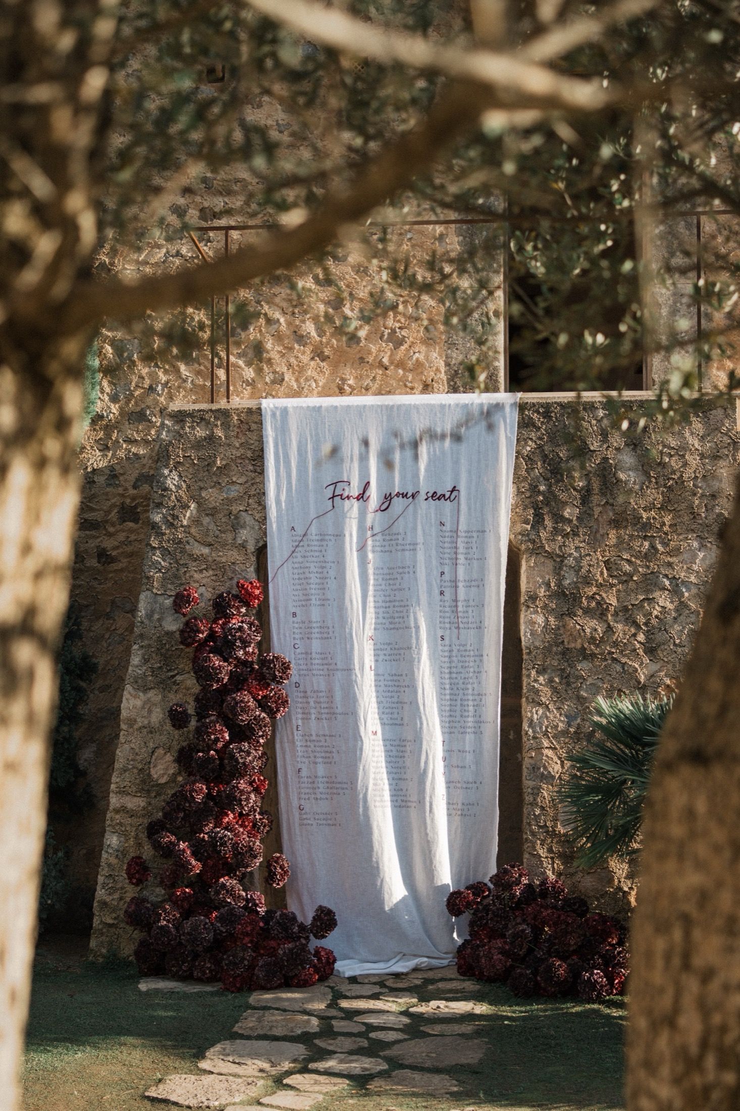 White sheer curtains hanging in a garden setting with red flowering bushes and tree trunks creating a natural backdrop.