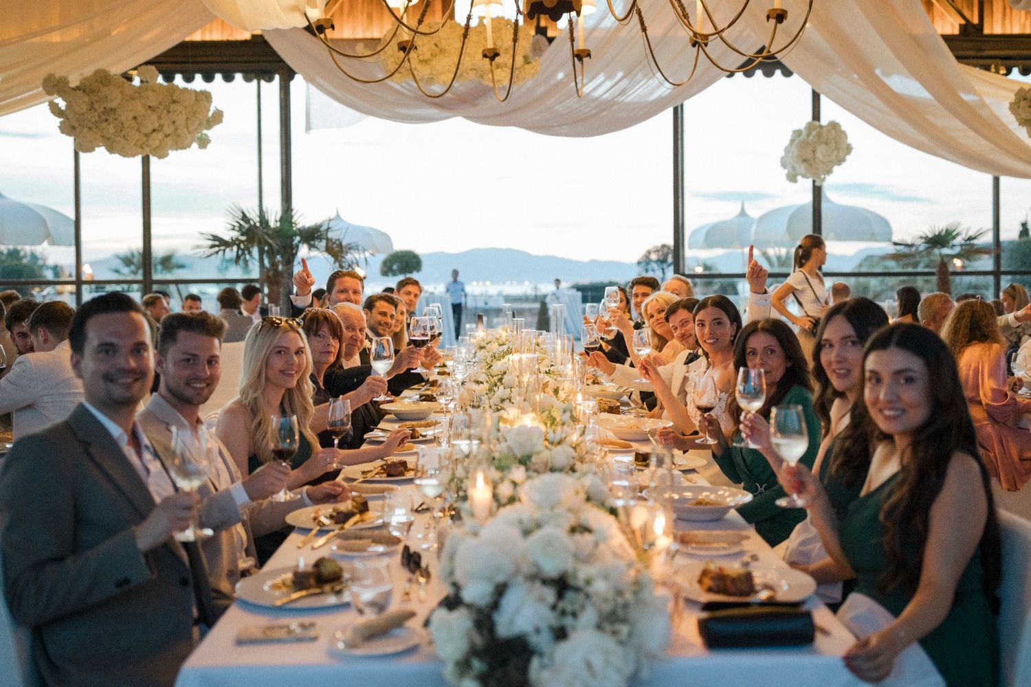Elegant wedding reception dinner with guests toasting at a long decorated table overlooking a scenic waterfront venue.