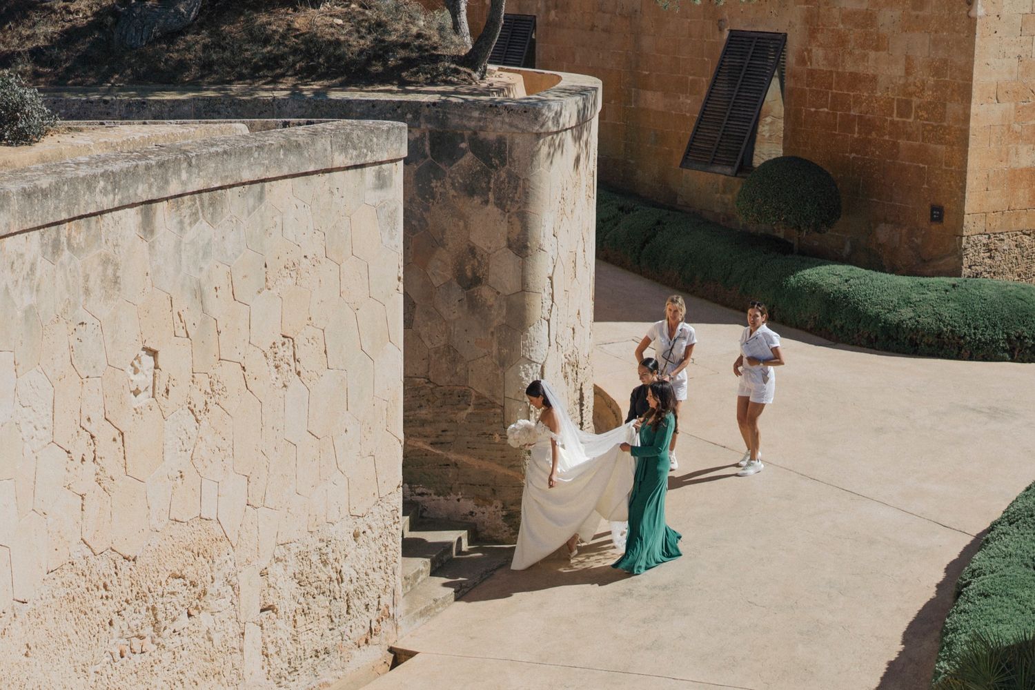 A bride in a white gown with attendants in green and white dresses at a historic stone fortress courtyard.