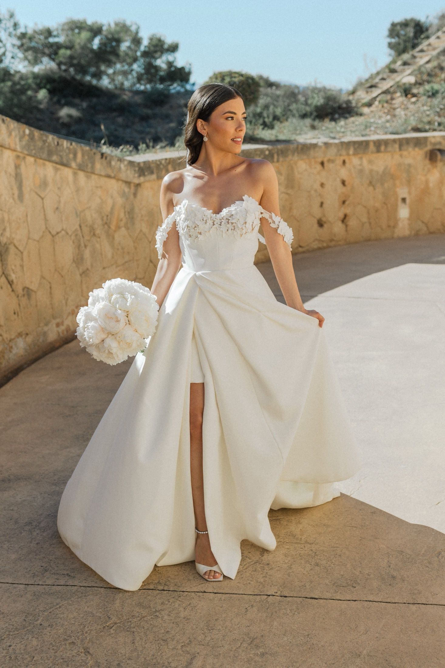 Bride in off-shoulder white gown with floral details and thigh slit holds bouquet outdoors in golden light.