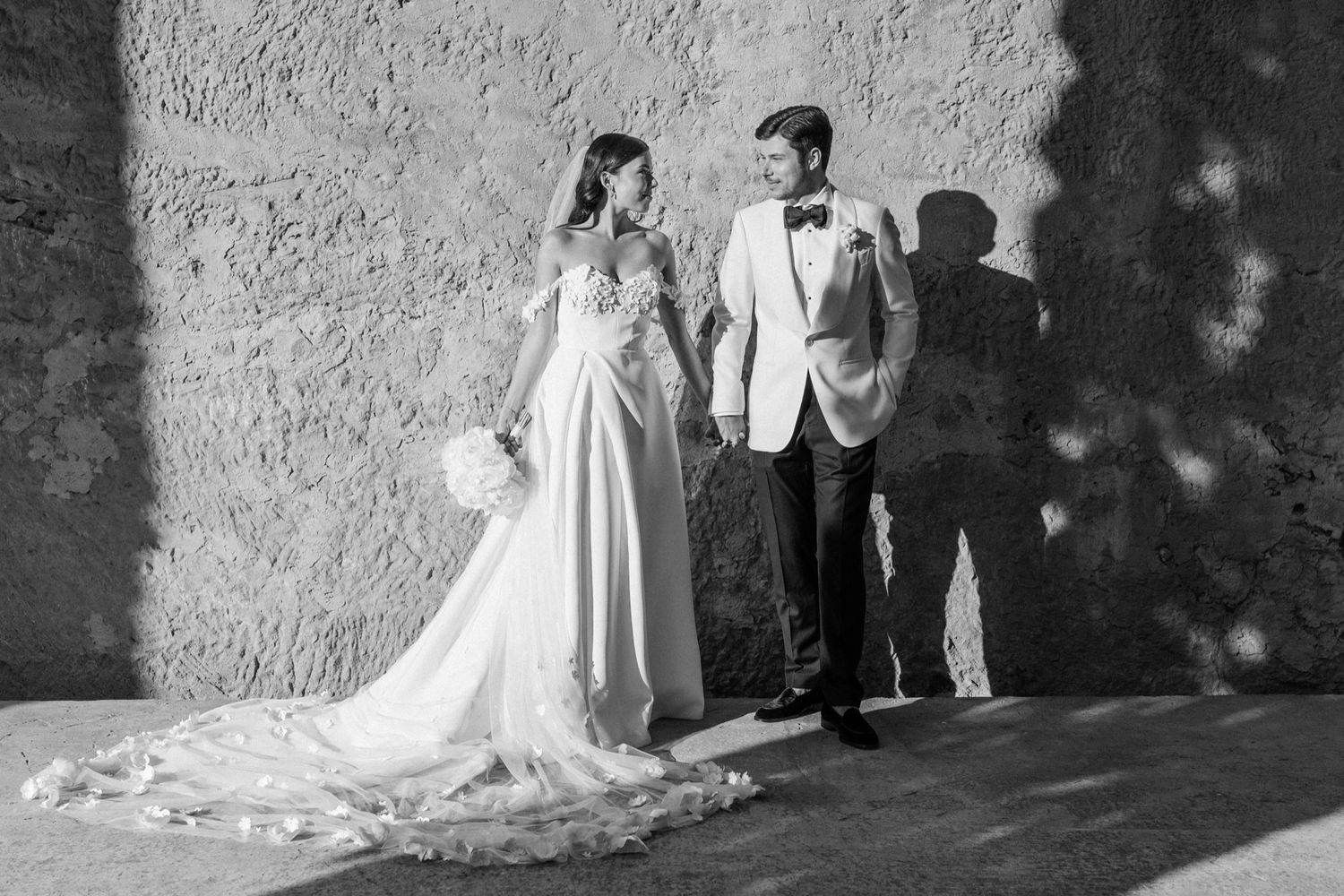 Bride and groom stand together in black and white photo against textured stone wall.
