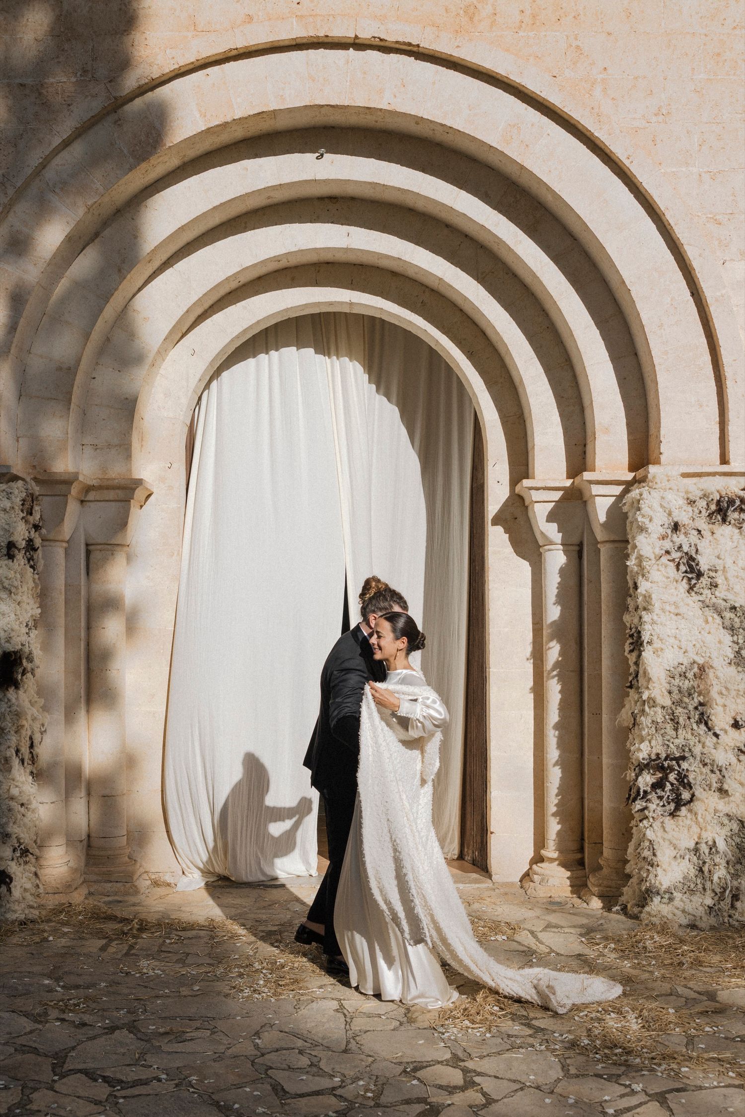Romantic wedding couple embracing under stone archways with white curtains and dramatic shadows.