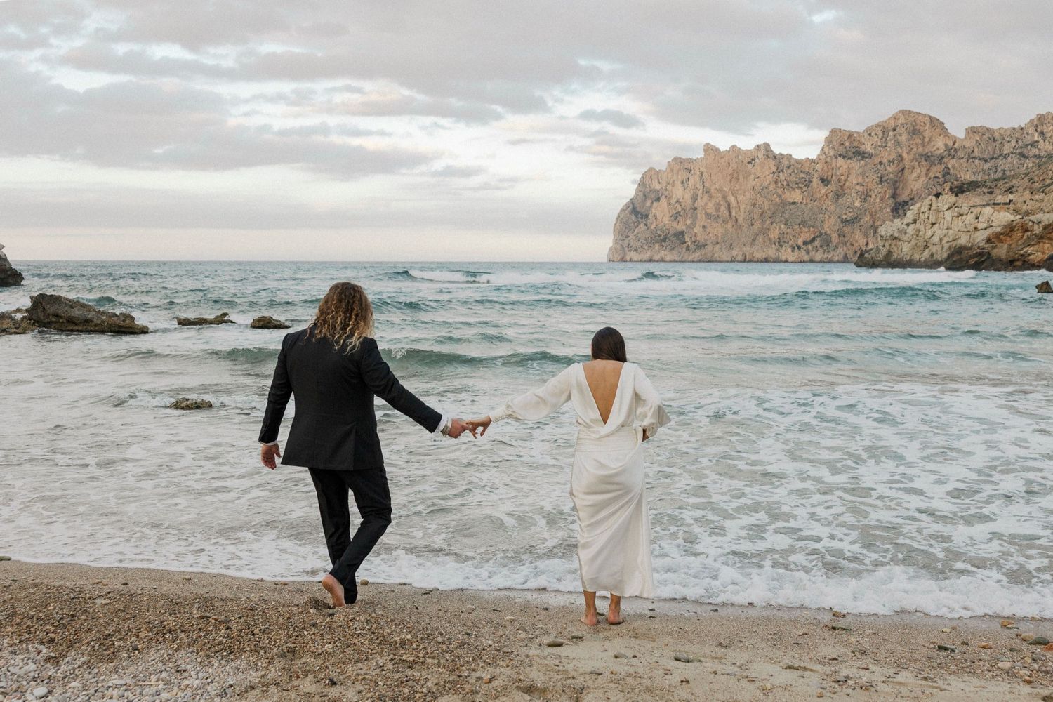 Two people strolling on a scenic beach with rocky cliffs and moody skies in the background.