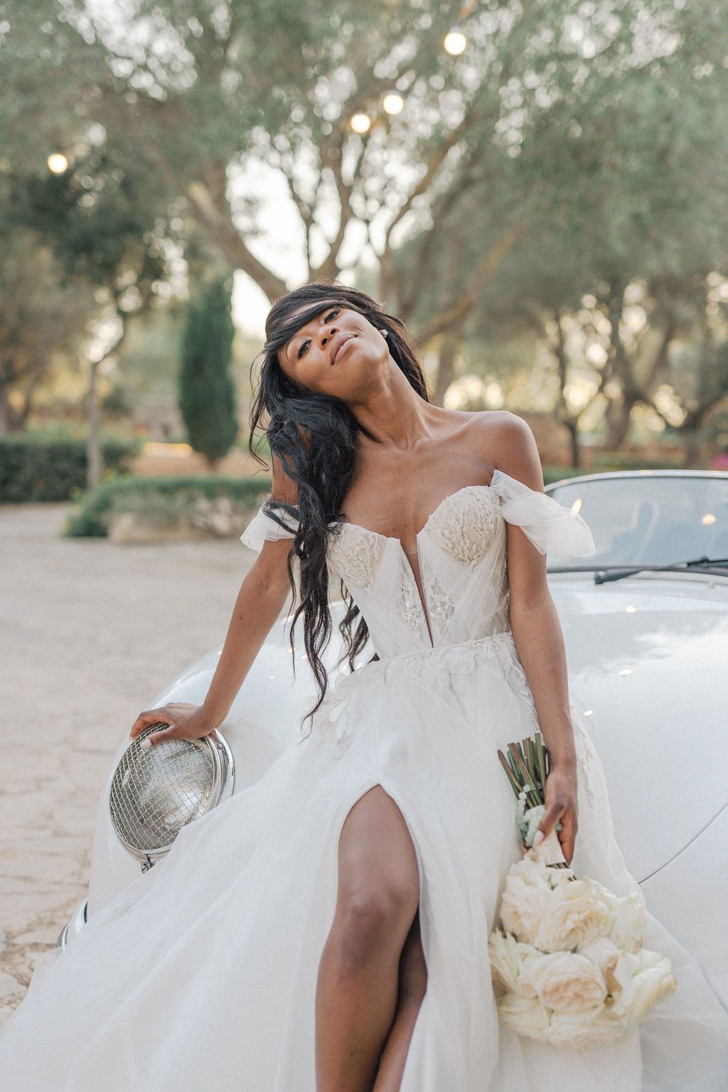 Bride in flowing white dress poses elegantly against a vintage car.