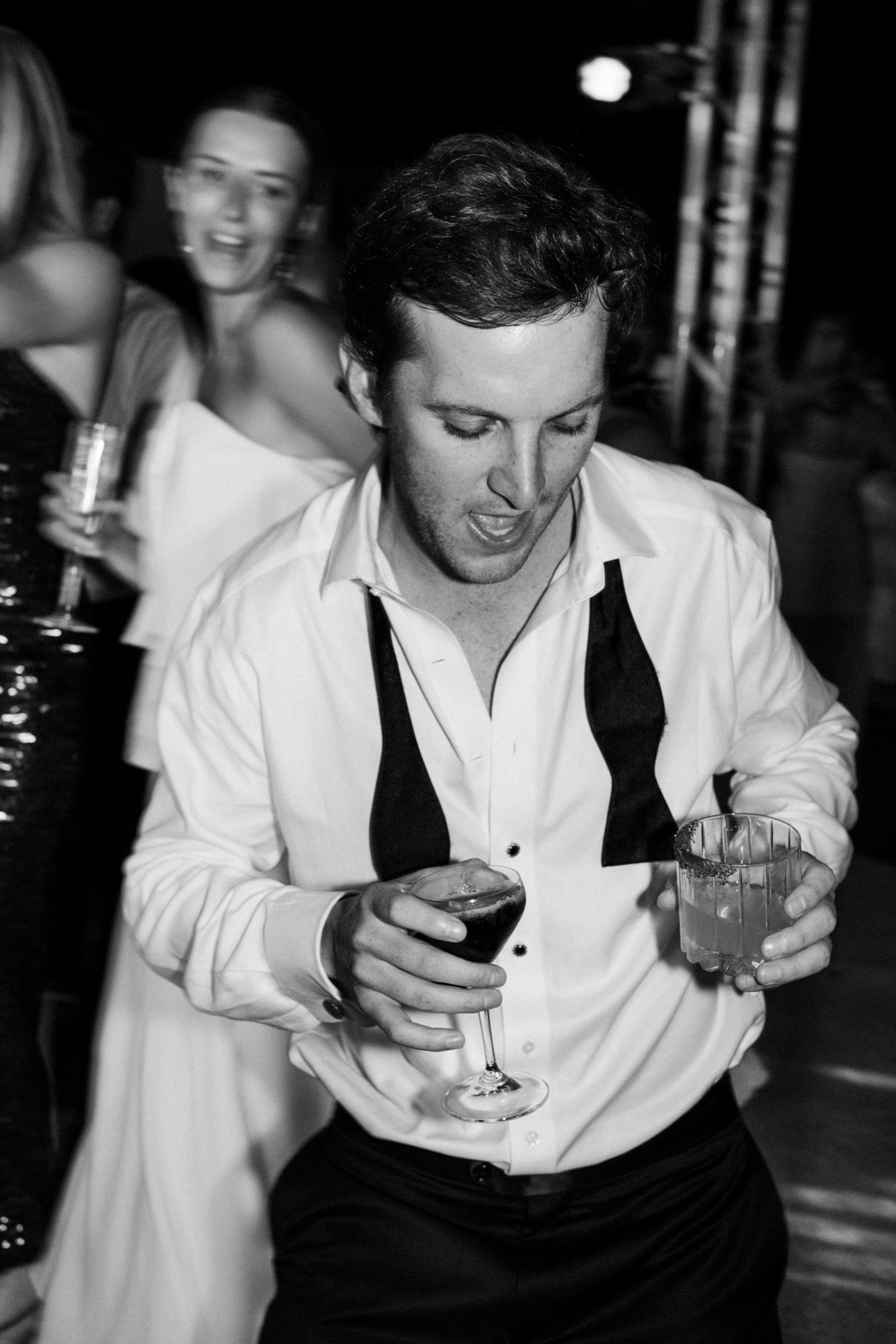 Party guest in white shirt and black tie dances while holding drinks at a nighttime celebration.
