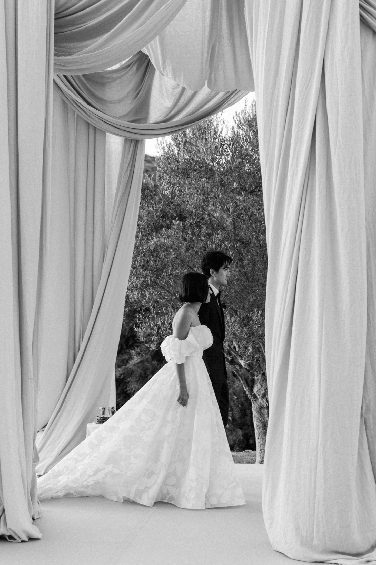 Elegant white fabric drapes frame a romantic outdoor wedding ceremony scene with silhouetted figures.