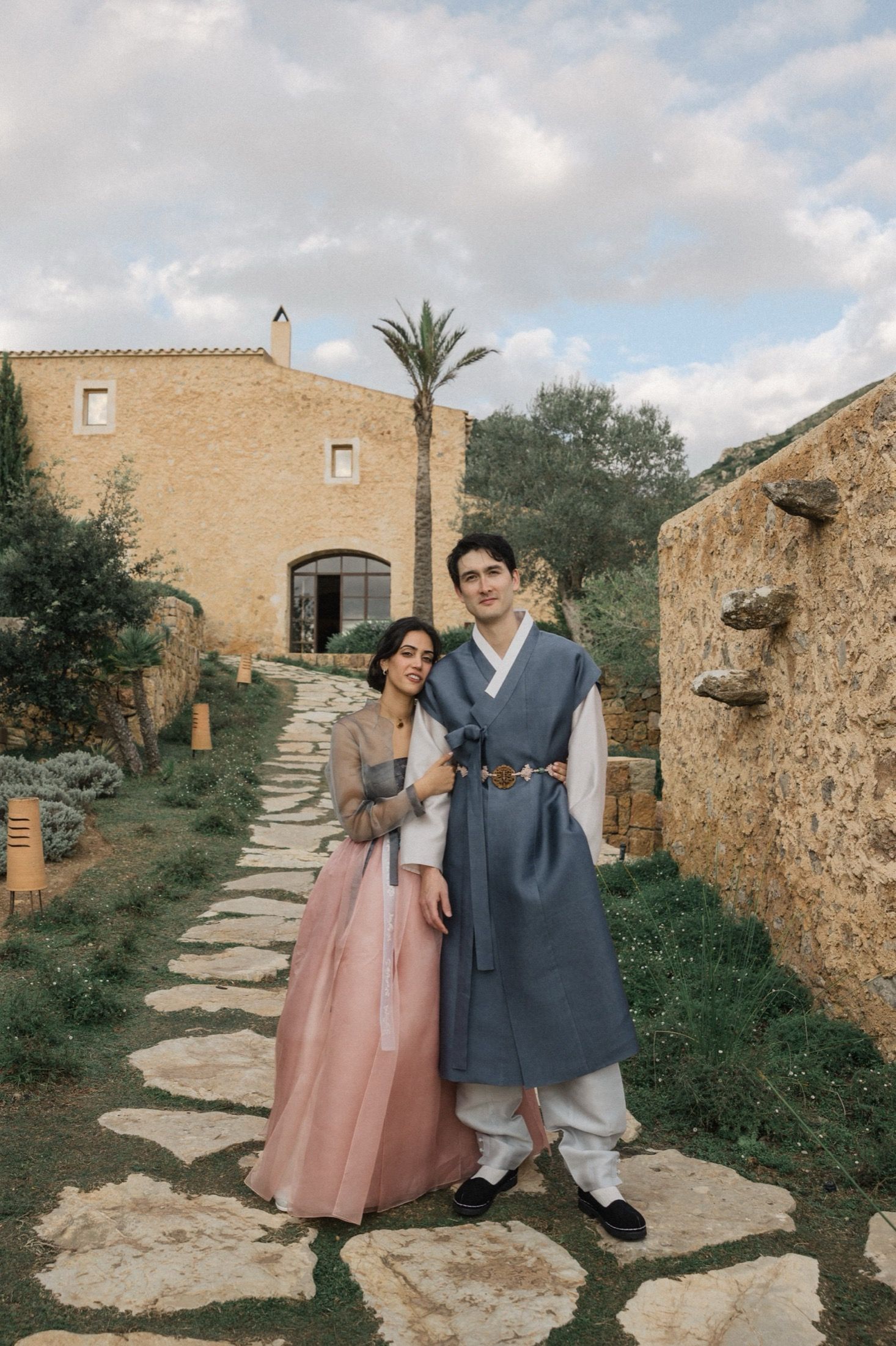 Two people in vintage style clothing pose on a stone path at a Mediterranean villa with stone walls and palm trees.