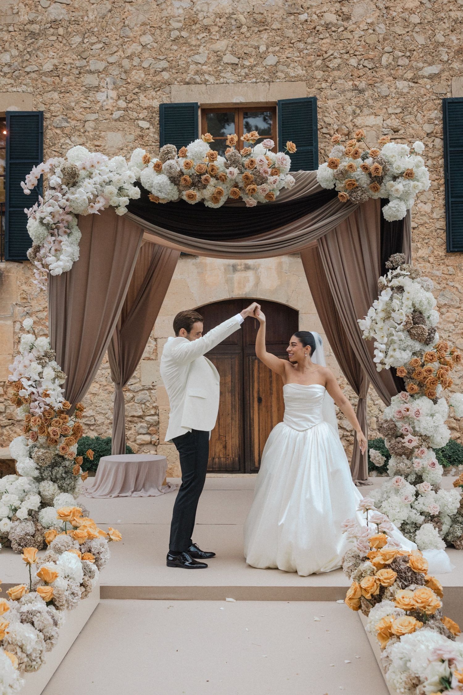 A couple dancing under an elegant wedding arch decorated with white and peach floral arrangements and taupe draping.