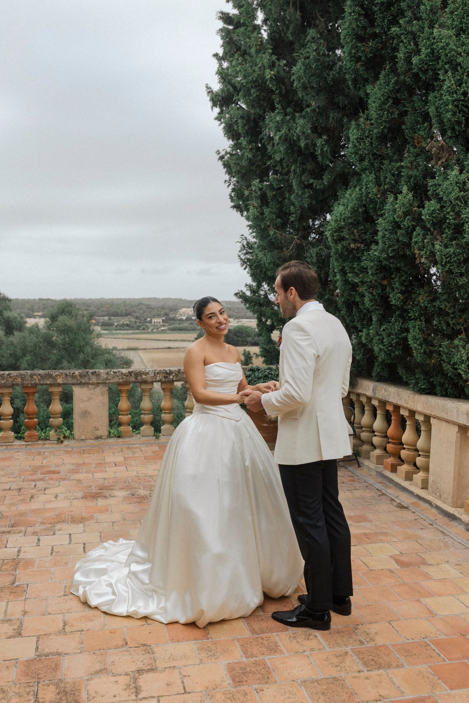 A couple shares an intimate moment on a terrace with stone balustrade and cypress trees overlooking a rural landscape.
