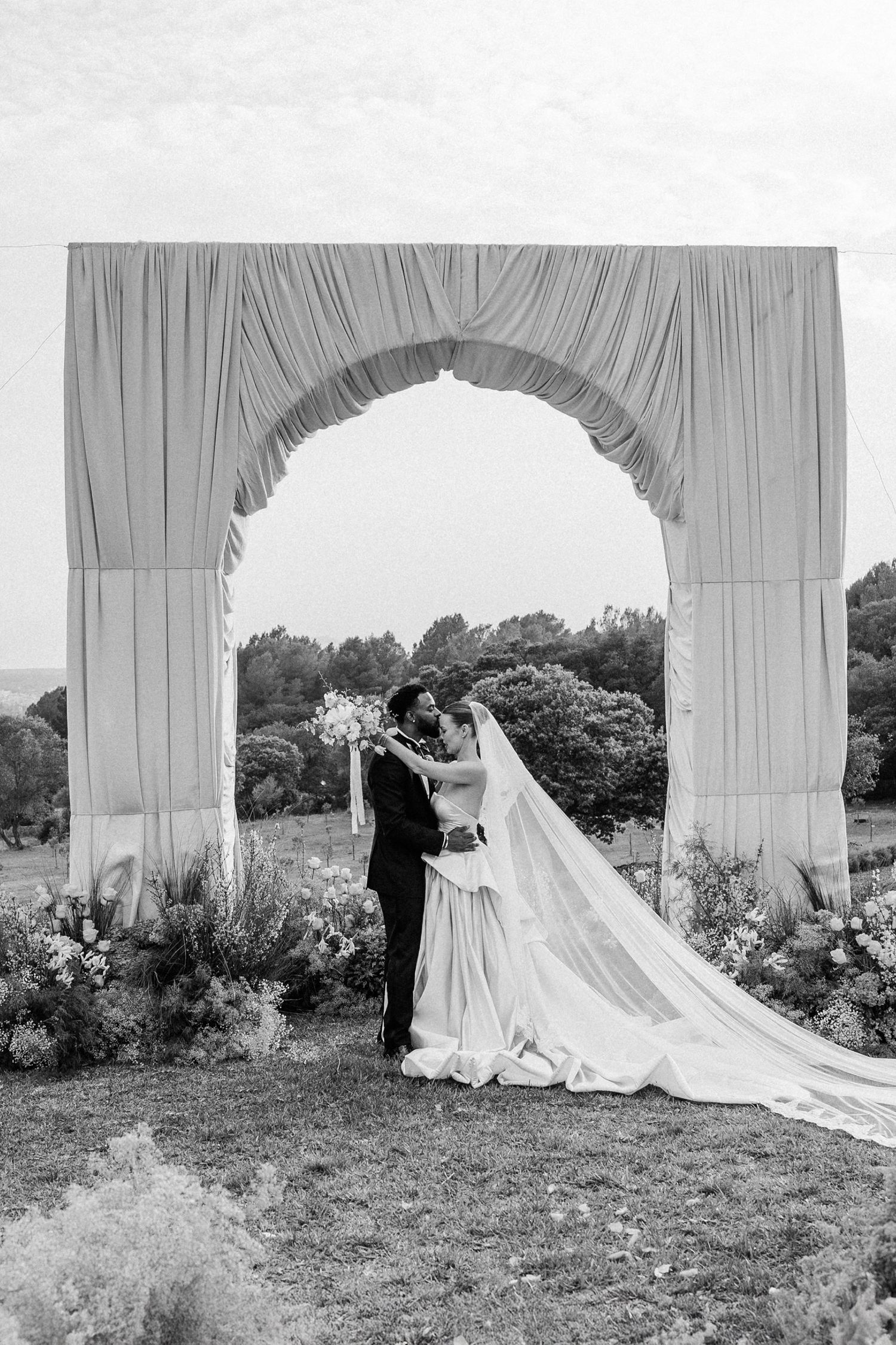 A black and white photo series shows a flowing wedding dress train and veil billowing in the wind through garden archways. Es Racó d'Artà