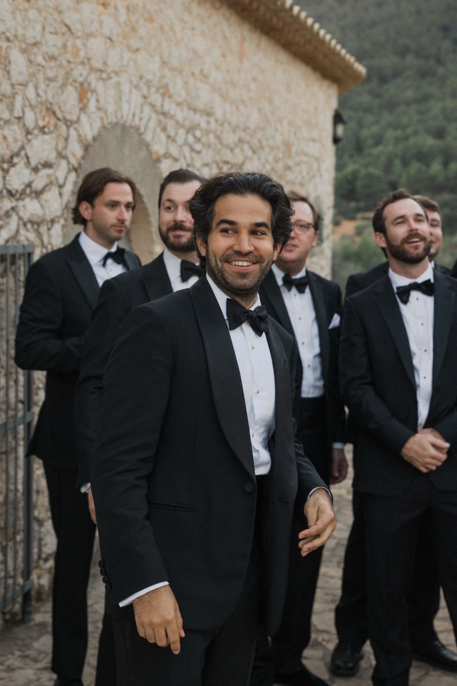 A group of groomsmen in black tuxedos and bow ties pose together outside a stone building.